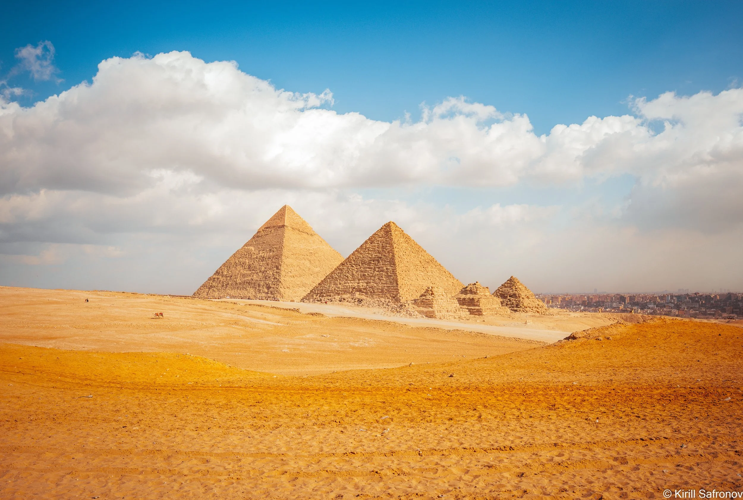 Photo of the Pyramids of Giza in the Egyptian desert under a clear blue sky with scattered clouds.