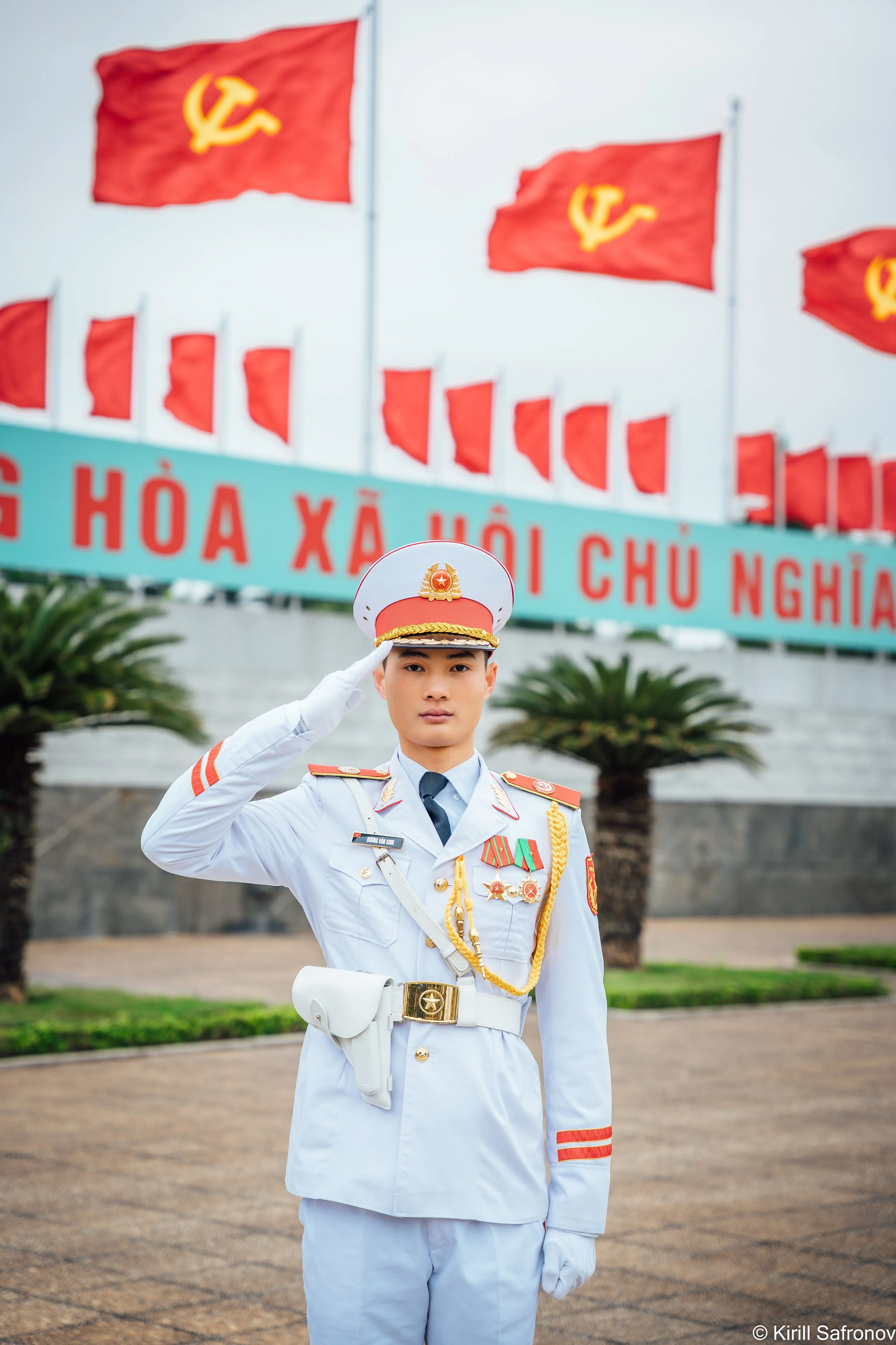 photo of a young man in a white military uniform saluting, wearing medals, standing before red flags and a Vietnamese sign