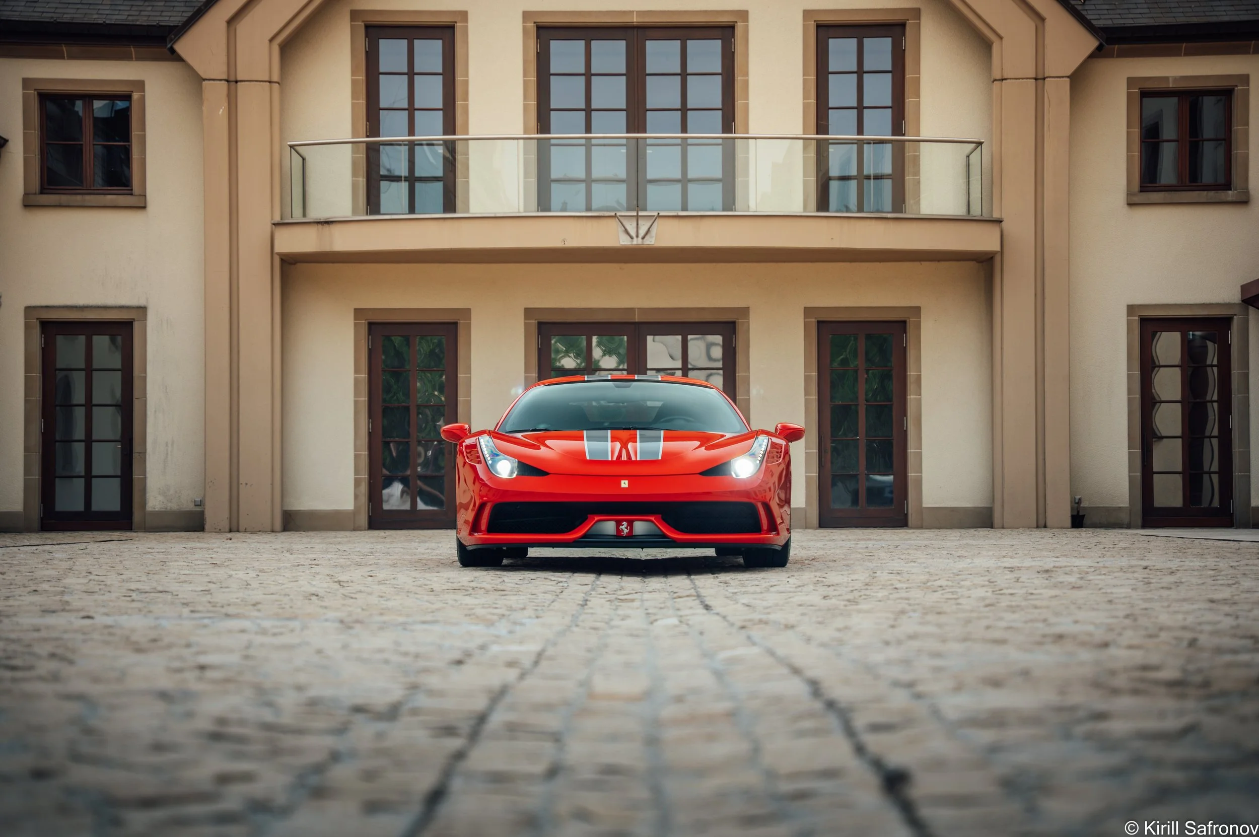 A red Ferrari 458 Speciale with white racing stripes on the hood, parked outside a modern luxury home featuring large glass windows, contemporary architecture, and a glass balcony.