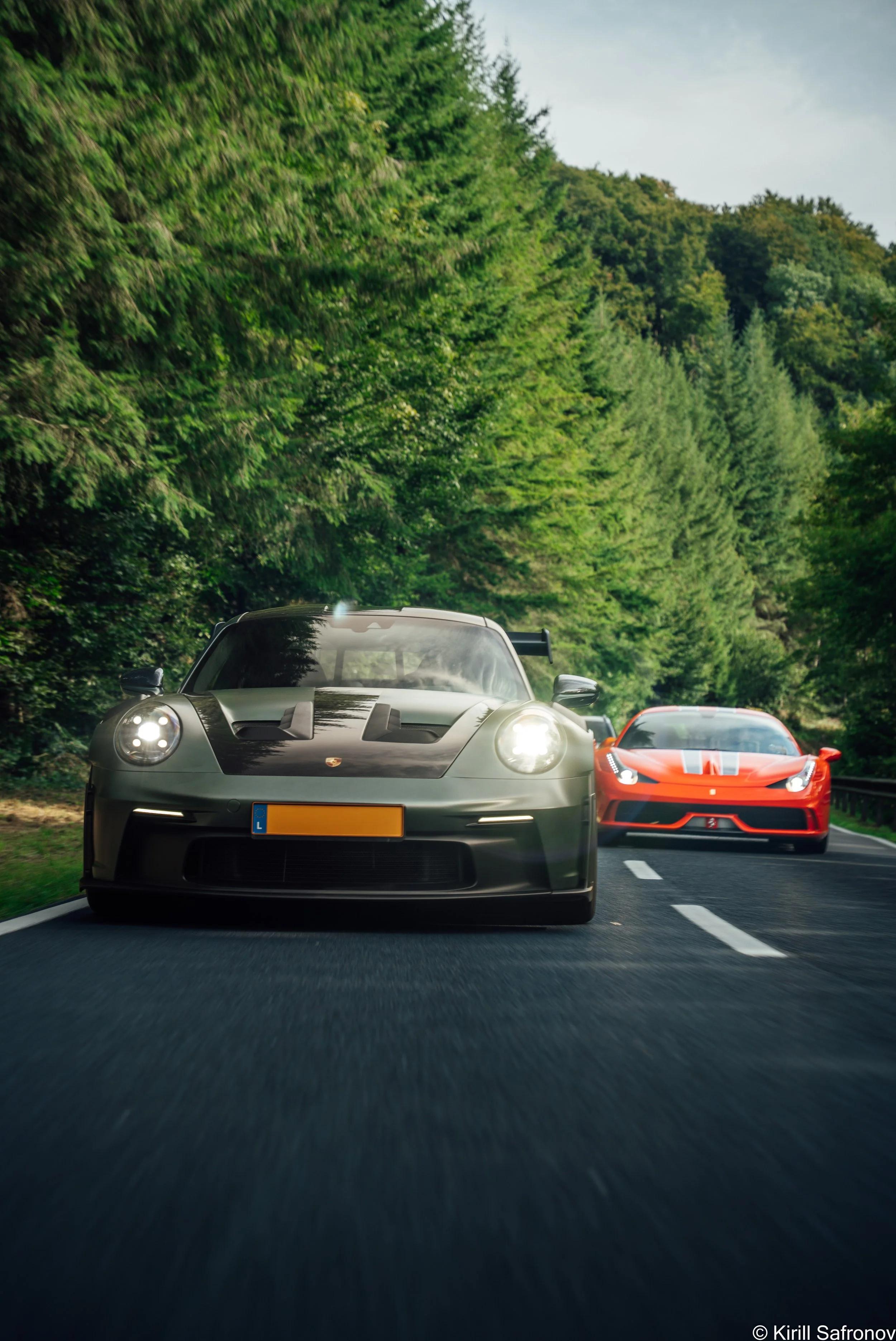 Dark green Porsche 911 GT3 RS and red Ferrari 458 Speciale driving together on a scenic forest road under a cloudy sky.