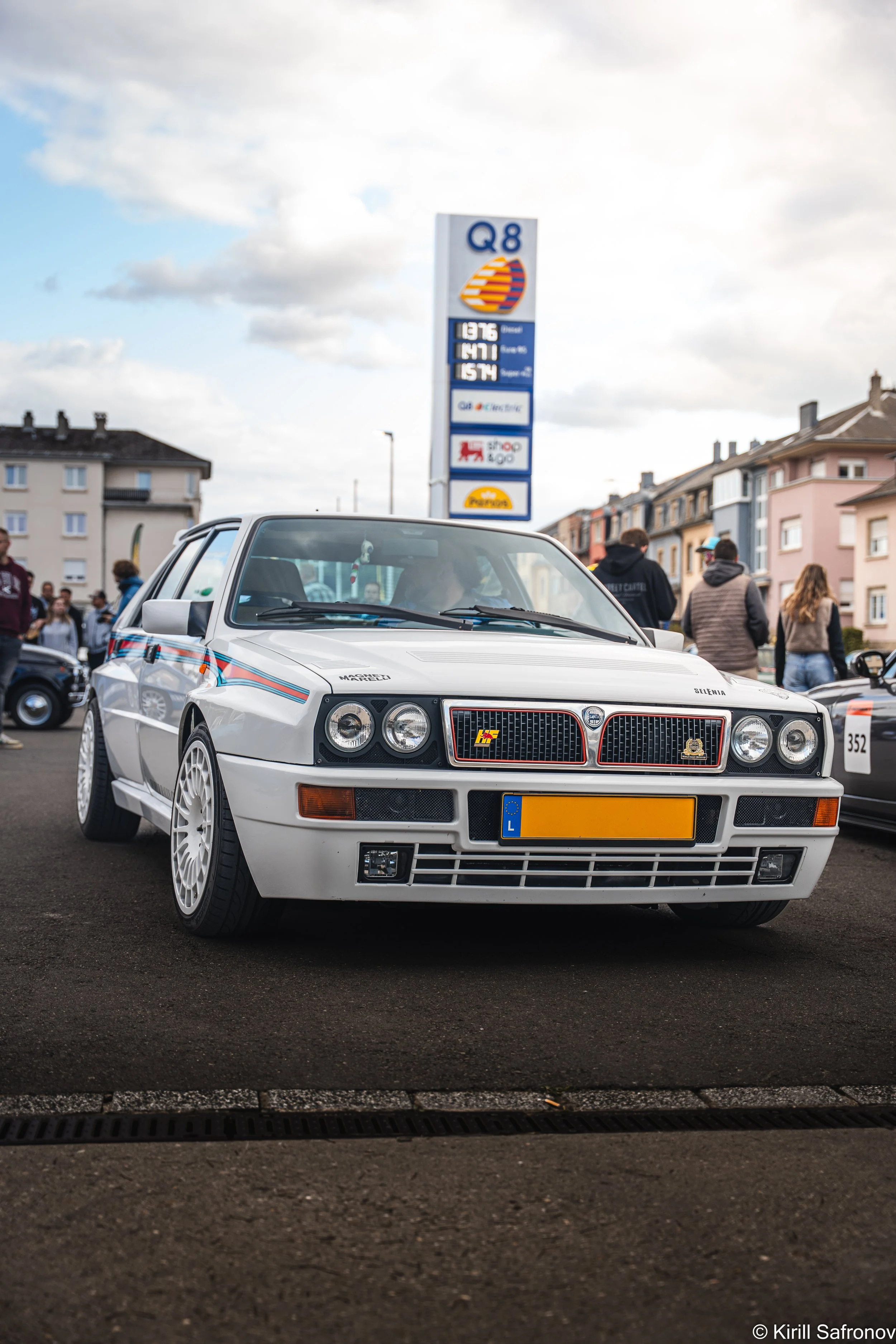 photo of a white vintage Lancia Delta Integrale rally car with racing decals, parked in an urban parking lot near residential buildings and a fuel price sign under a partly cloudy sky.