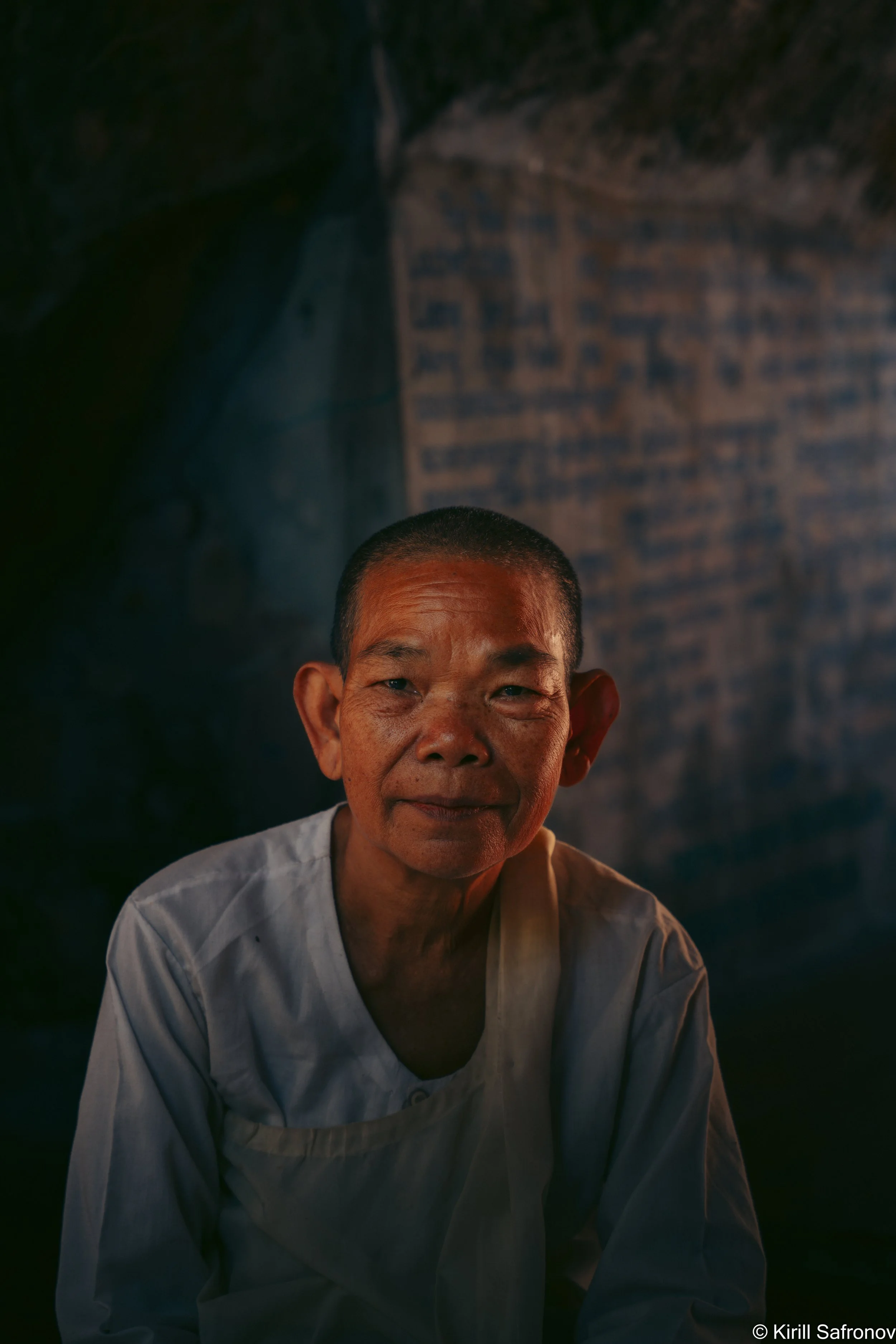 photo of an elderly Cambodian female monk with a gentle smile, dressed in a white robe, standing in a calm dimly lit interior against a simple wall background.