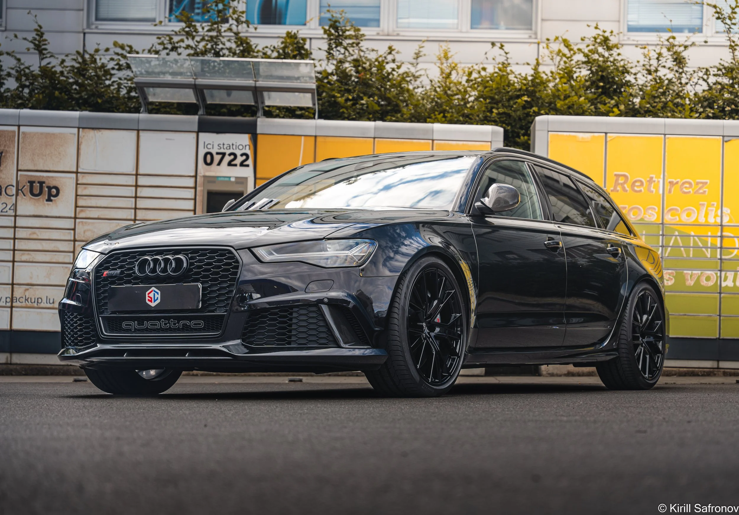 Photo of a black Audi Quattro RS6 C7 driving past a bus stop with yellow signs and a green hedge in the background.