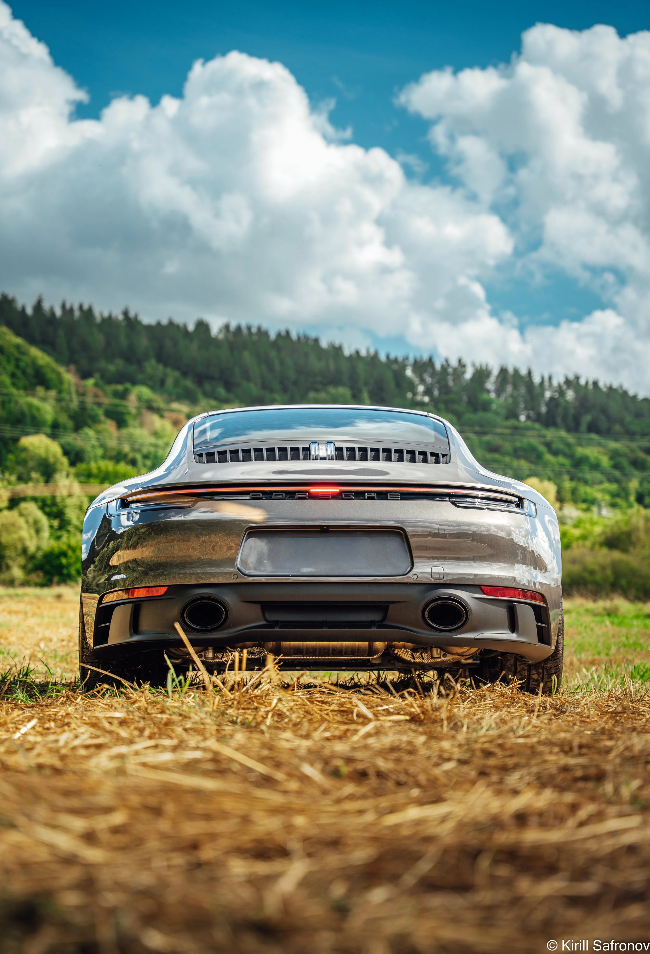 Rear view photo of a champagne Porsche 911 Carrera GTS parked in a countryside field, with wooded hills and a dramatic cloudy sky in the background