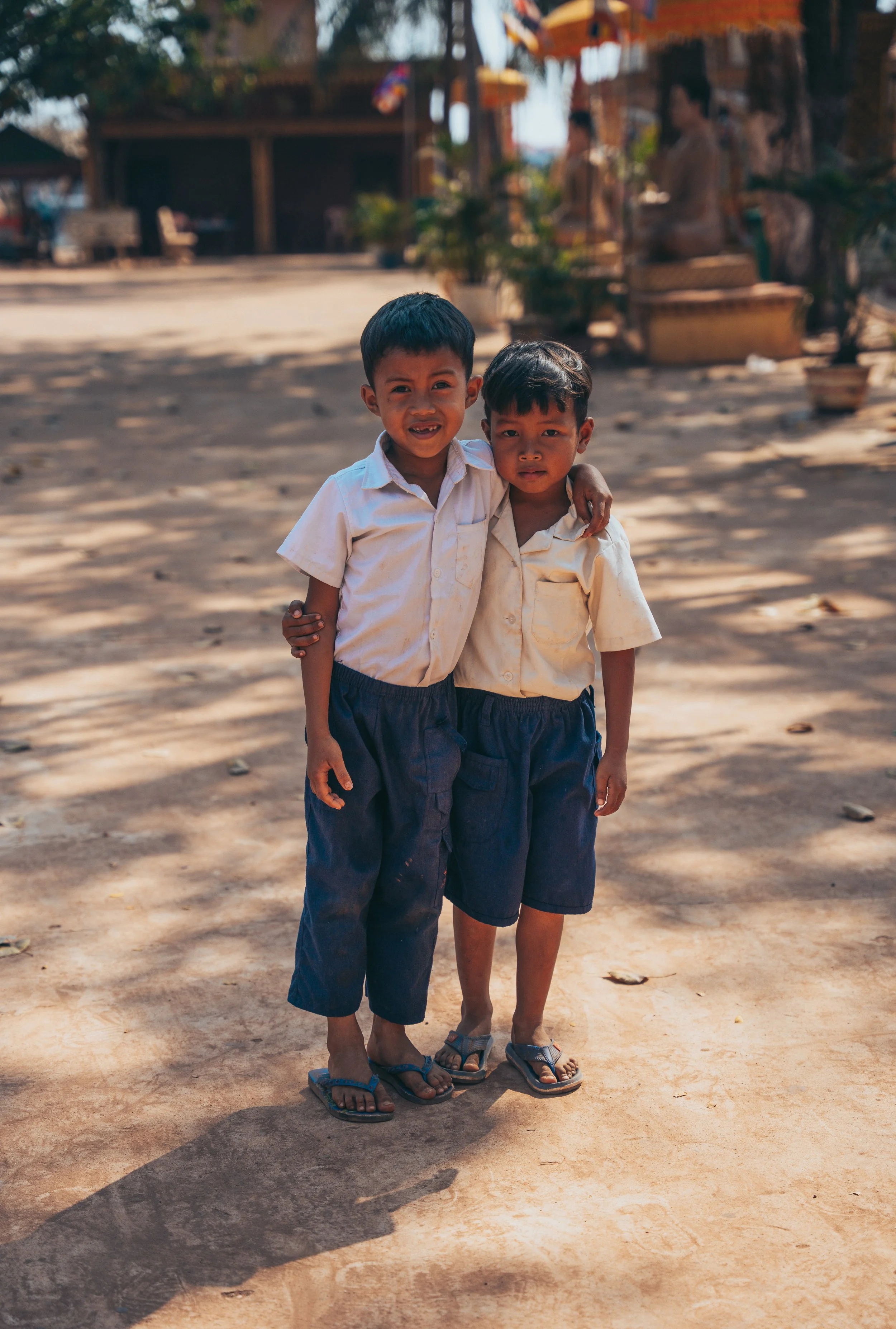 Photo of two smiling young boys wearing white and blue school uniforms and sandals, standing in a dirt courtyard under a tree, with school buildings in the background.