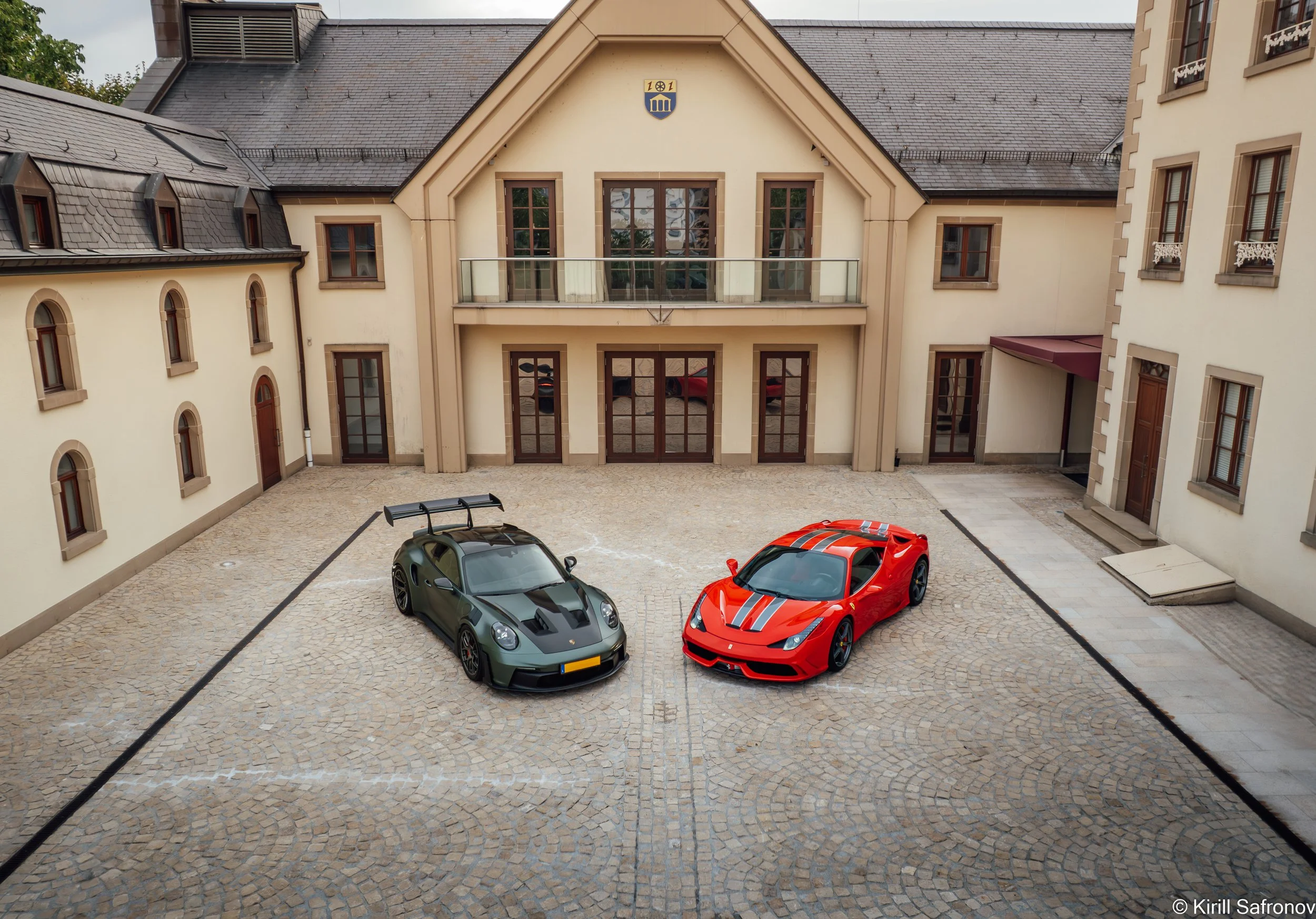 Dark green Porsche 911 GT3 RS and red Ferrari 458 Speciale parked in a paved courtyard in front of a beige historic building with large windows and a slate roof.