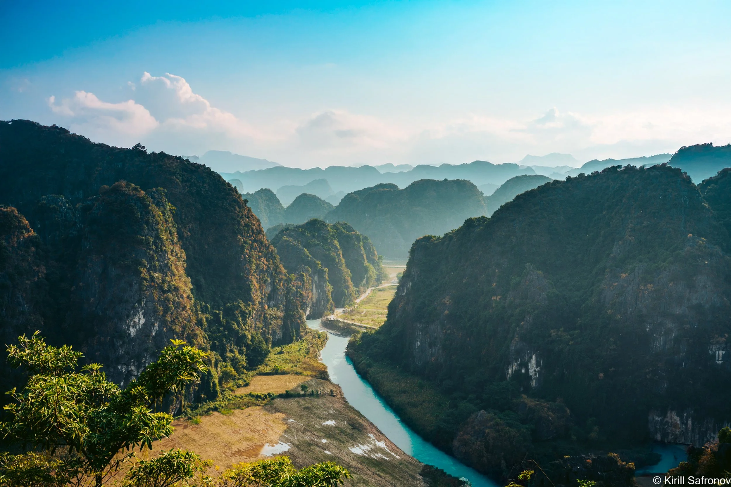 Photo of Ninh Binh, Vietnam, featuring lush green mountains, a winding blue river, and a clear blue sky with soft clouds. This peaceful landscape captures the natural beauty of northern Vietnam, making it ideal for travel photography, nature photogra