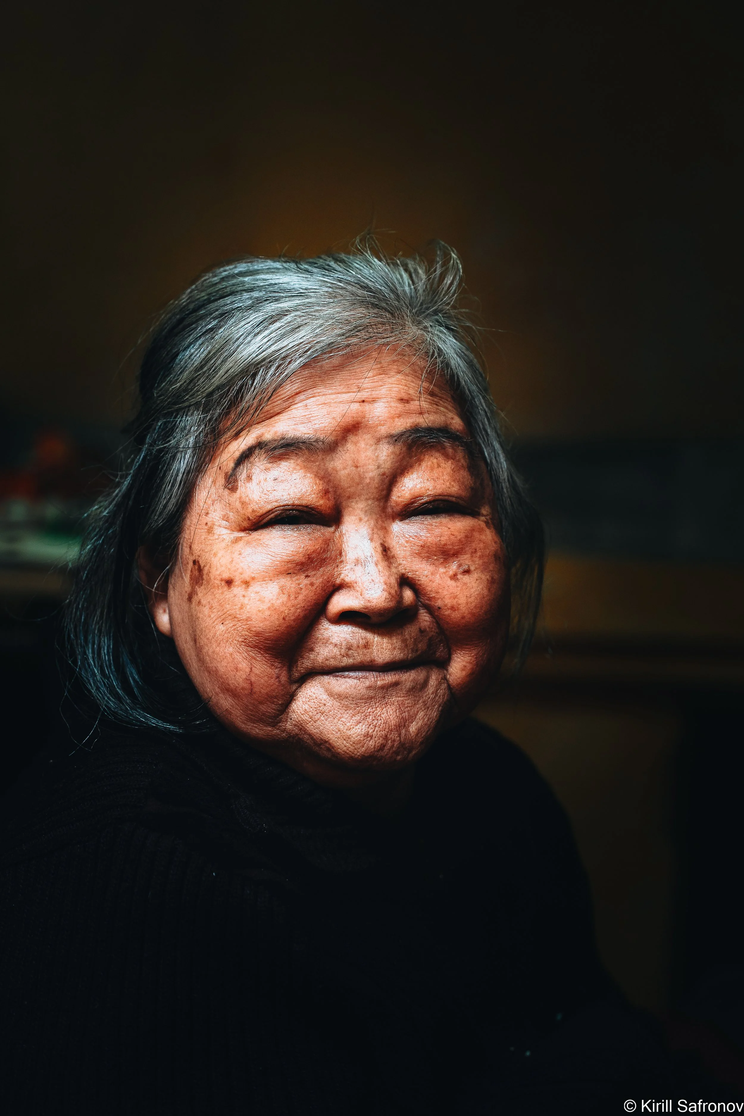 photo of a smiling elderly Vietnamese woman with gray hair wearing a black sweater against a dark background, creating a warm and authentic portrait with strong contrast and emotional expression.