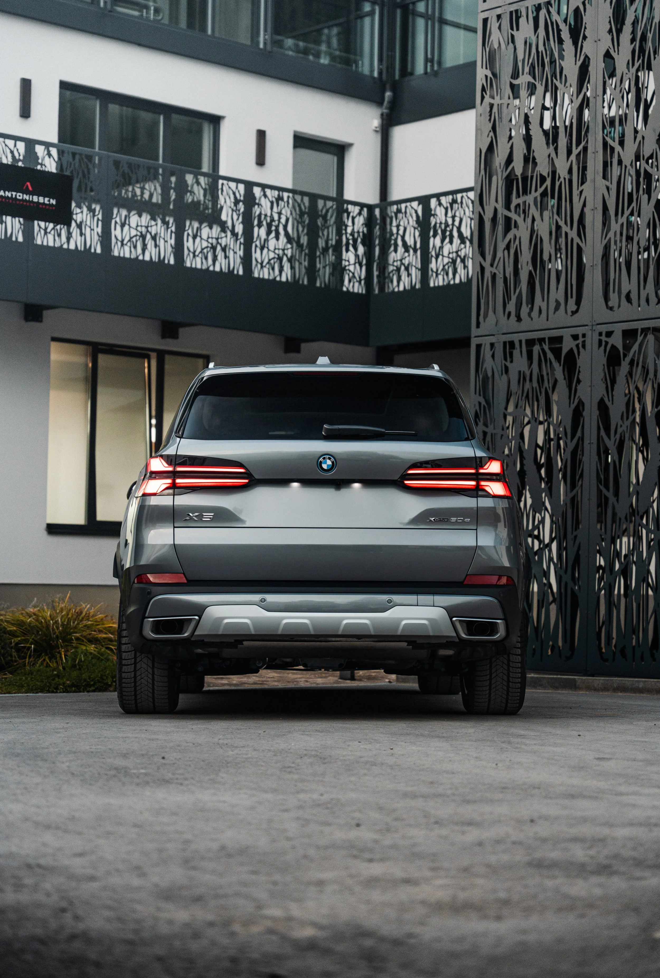 Photo of a dark gray BMW X5 rear view, parked outside a modern residential building with large windows and decorative metal balconies.