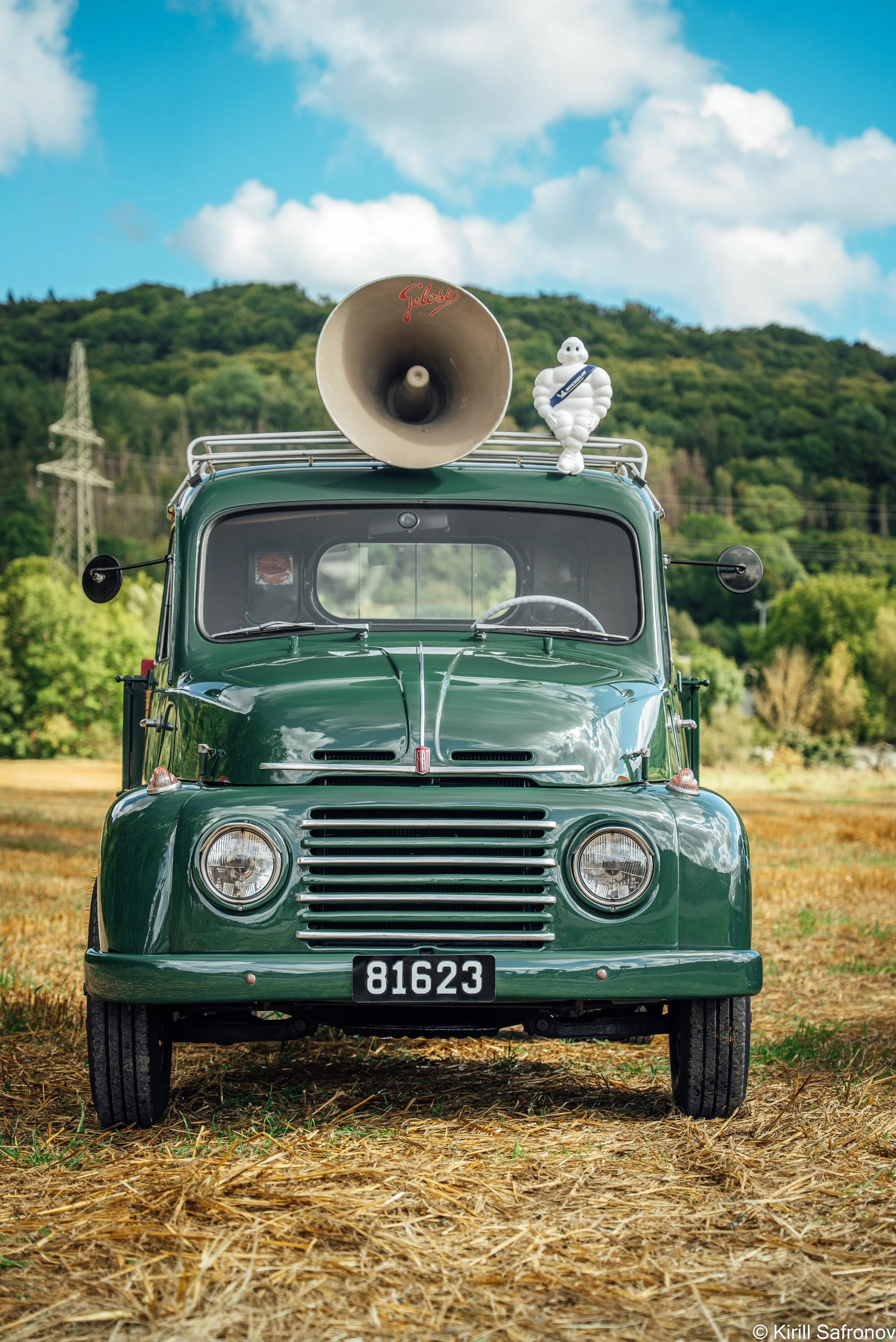 Photo of a dark green 1995 Fiat 615 vintage truck featuring a Michelin advertising sign, roof loudspeaker, and Michelin Man figure, with a blue sky, clouds, and green hills in the background.