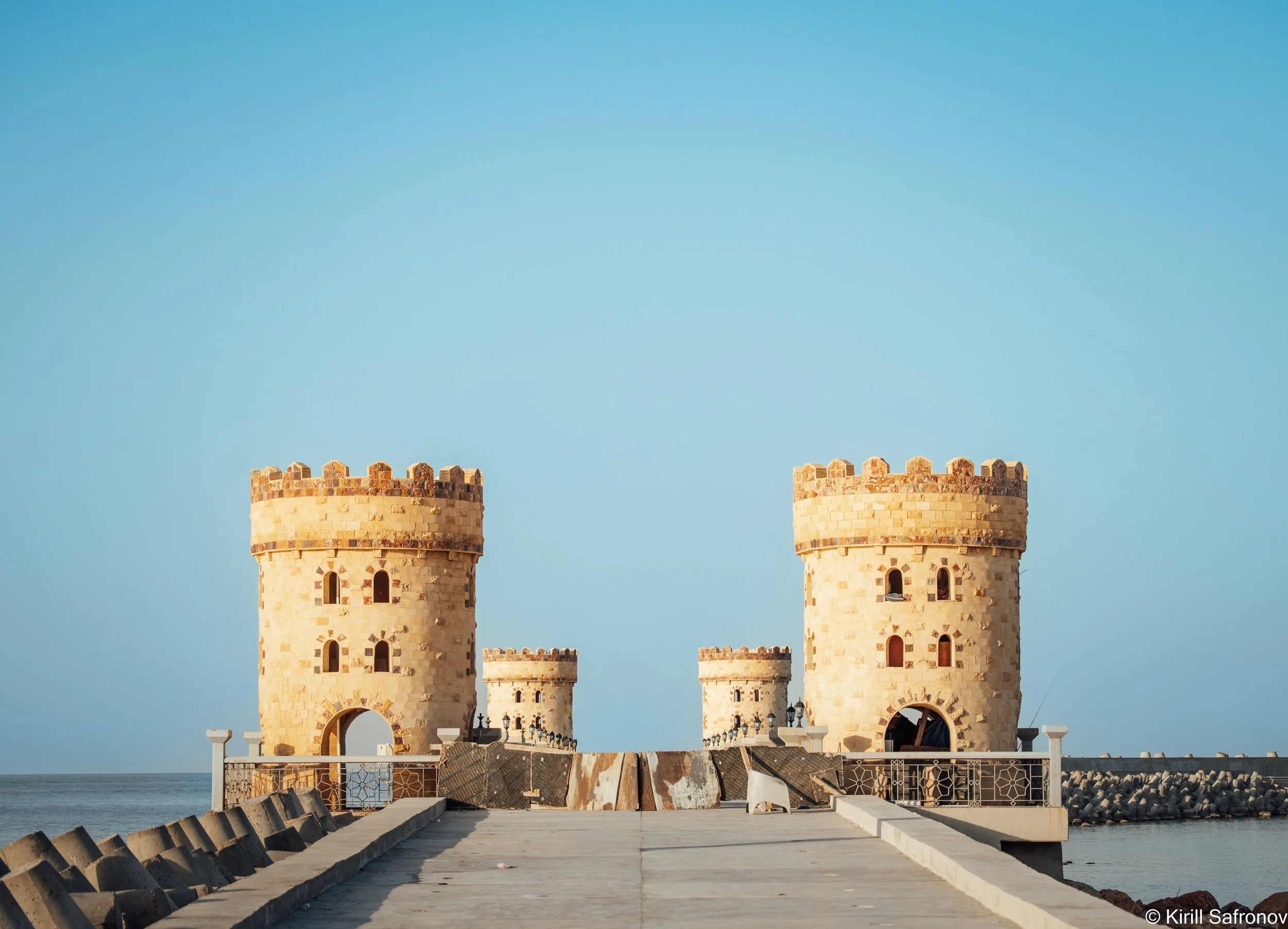 Photo of the Lighthouse of Alexandria in Egypt, showing two beige stone towers with arched windows, a concrete walkway, the Mediterranean Sea, and a clear blue sky.