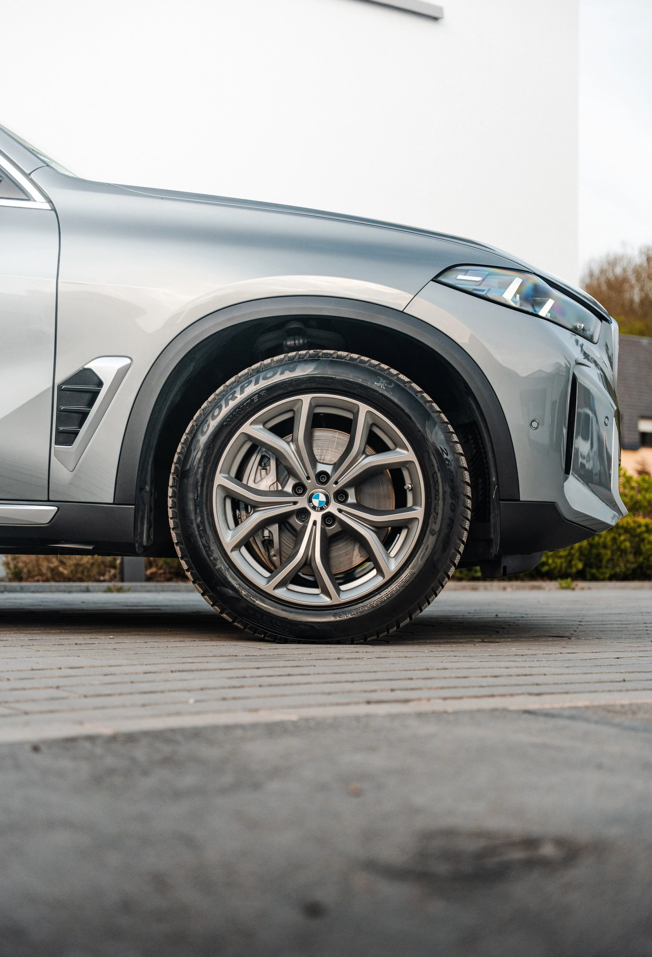 Photo of a dark gray BMW X5 parked on a wooden path in daylight, showing a partial front view, front wheel, and left side of the luxury SUV.