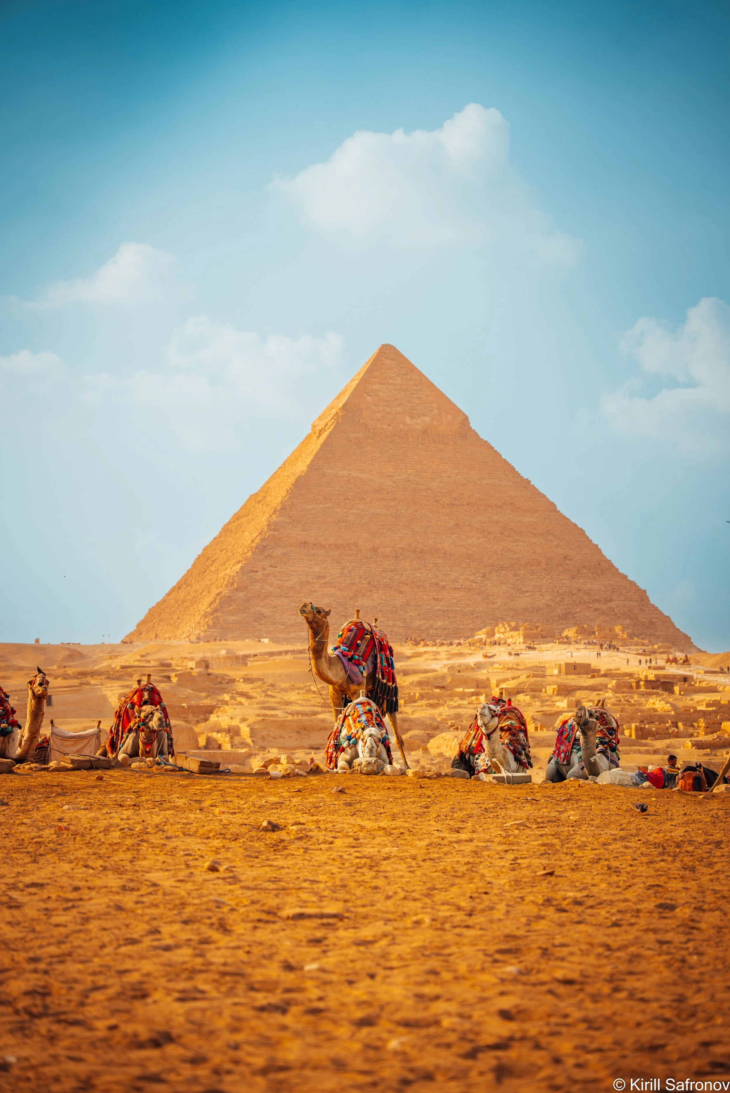 Photo of the Giza pyramids with camels and tourists in the foreground under a blue sky with scattered clouds.