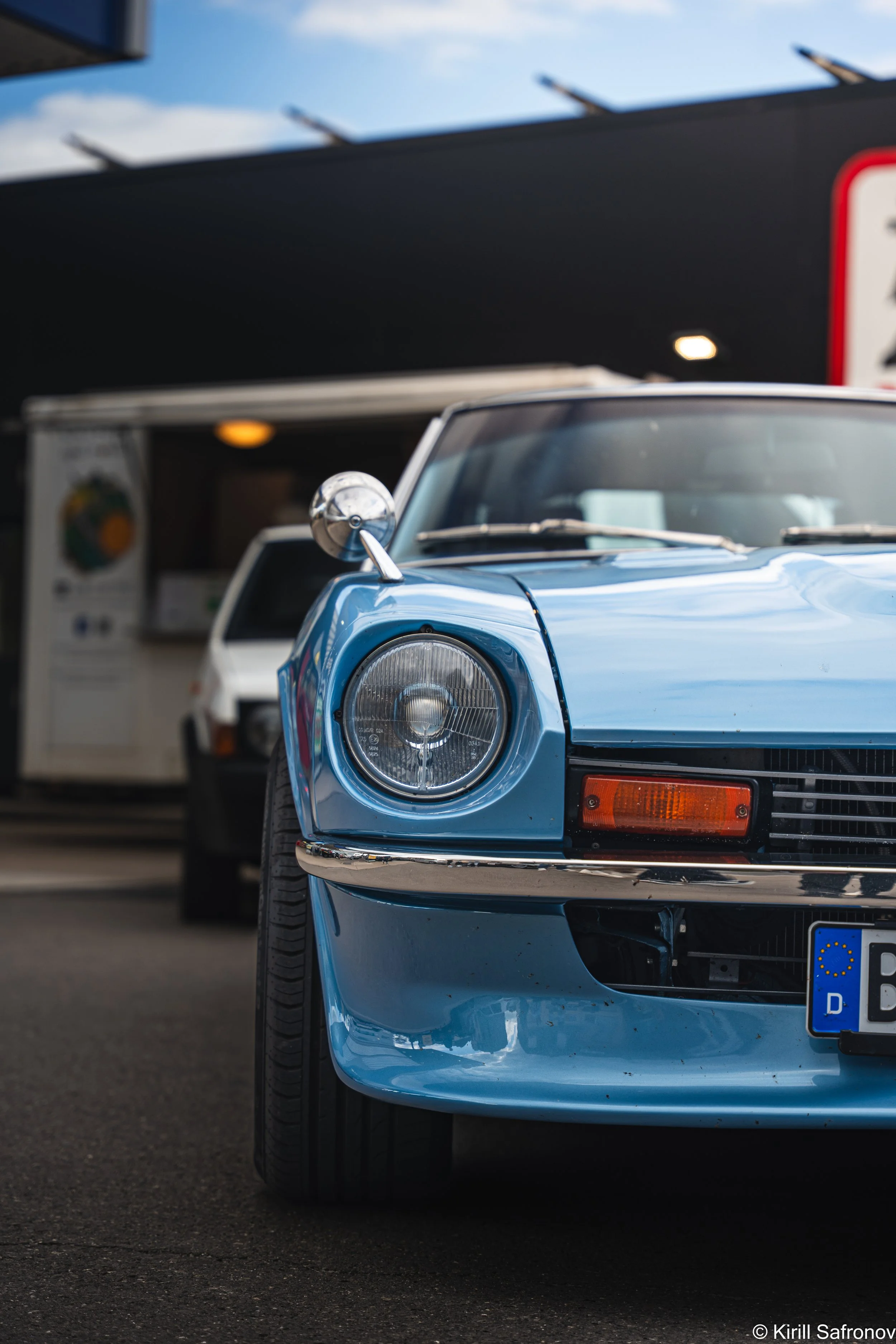 Photo of a classic blue 1976 Datsun 280Z sports car with round headlights, parked on a road with other vehicles in the background under a partly cloudy sky.