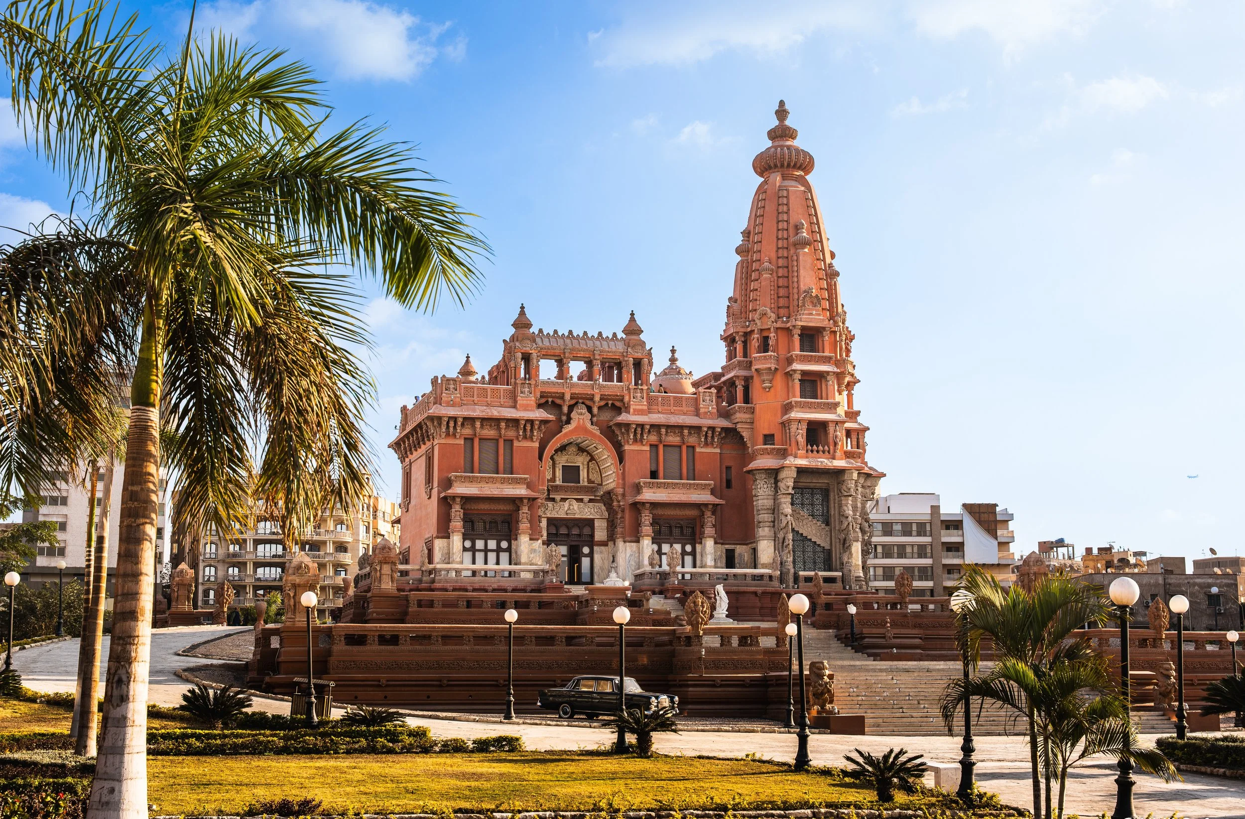 Photo of Baron Empain Palace, a historic Indian-inspired architectural building featuring a pointed tower, palm trees, and streetlights in an urban cityscape.