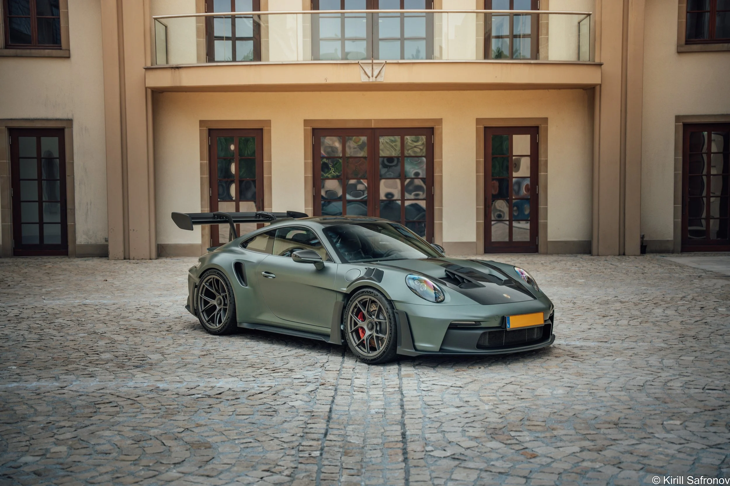 Dark green Porsche 911 GT3 RS parked on a cobblestone square in front of a modern building with glass windows and wooden doors.