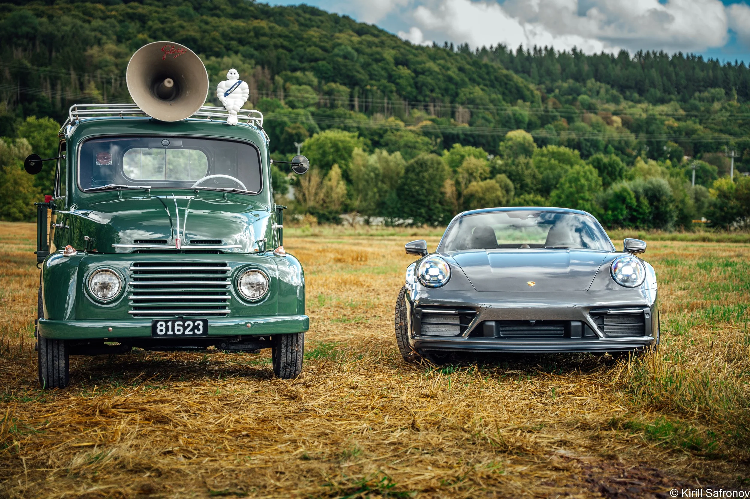 Photo of a champagne-colored Porsche 911 Carrera GTS parked beside a dark green 1995 Fiat 615 truck with a Michelin advertising sign, set in an open field with a forested mountain in the background.
