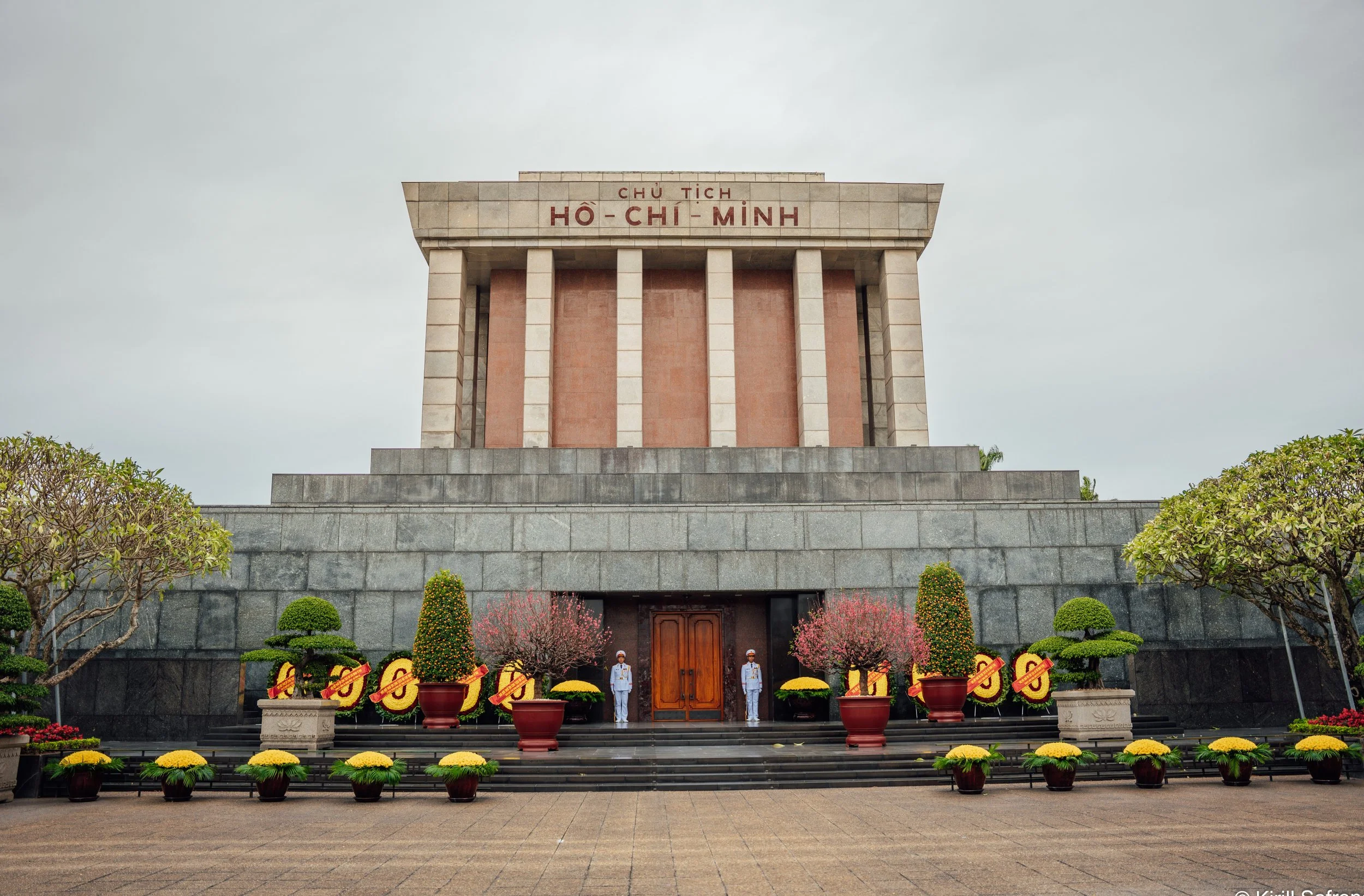 photo of an official building in Hanoi decorated with flowers, with two uniformed soldiers standing guard at the main entrance and a Vietnamese flag flying on the facade.