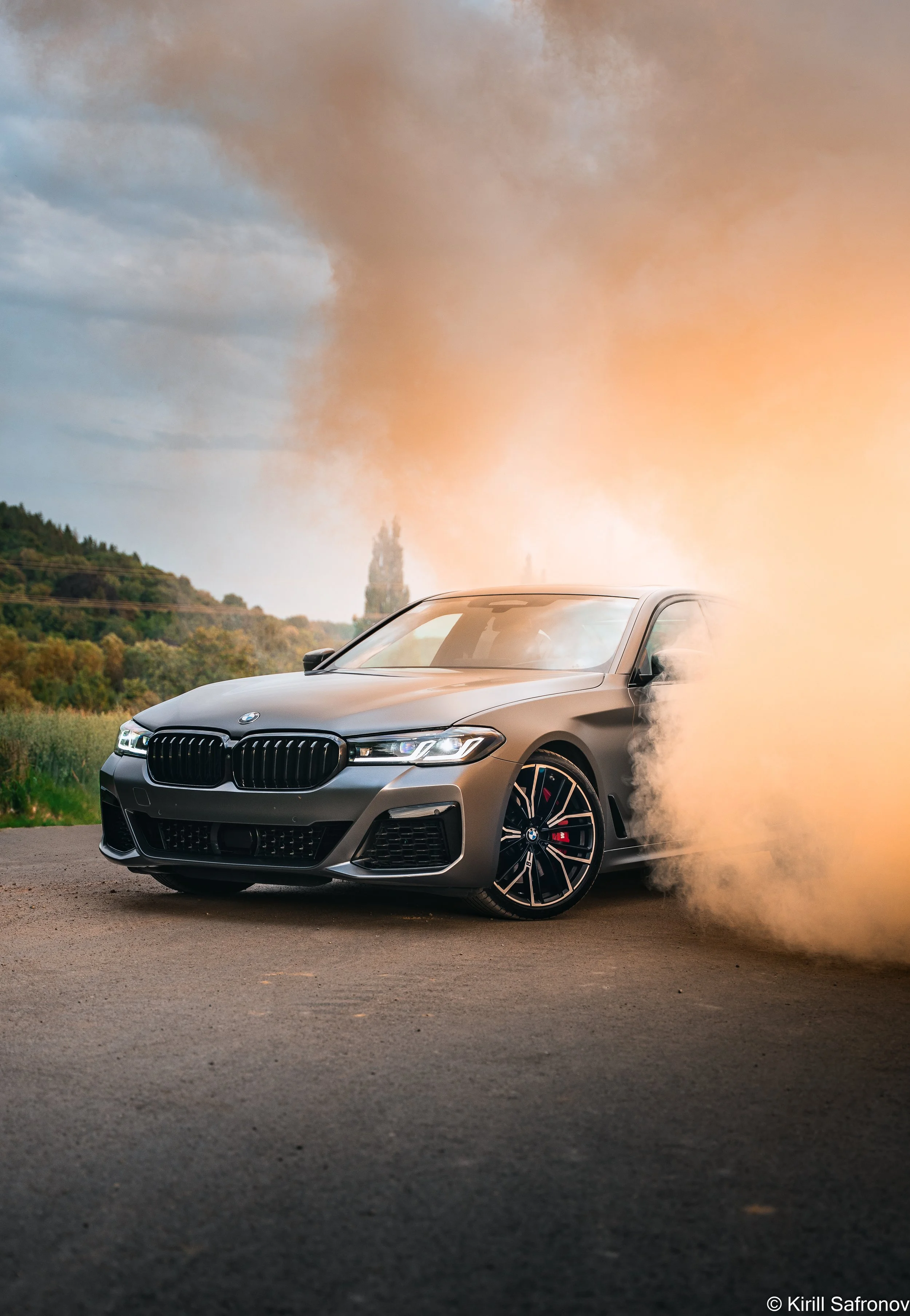 Photo of a grey BMW M550i driving on a rural road, kicking up dust, with a cloudy sky and countryside landscape in the background.