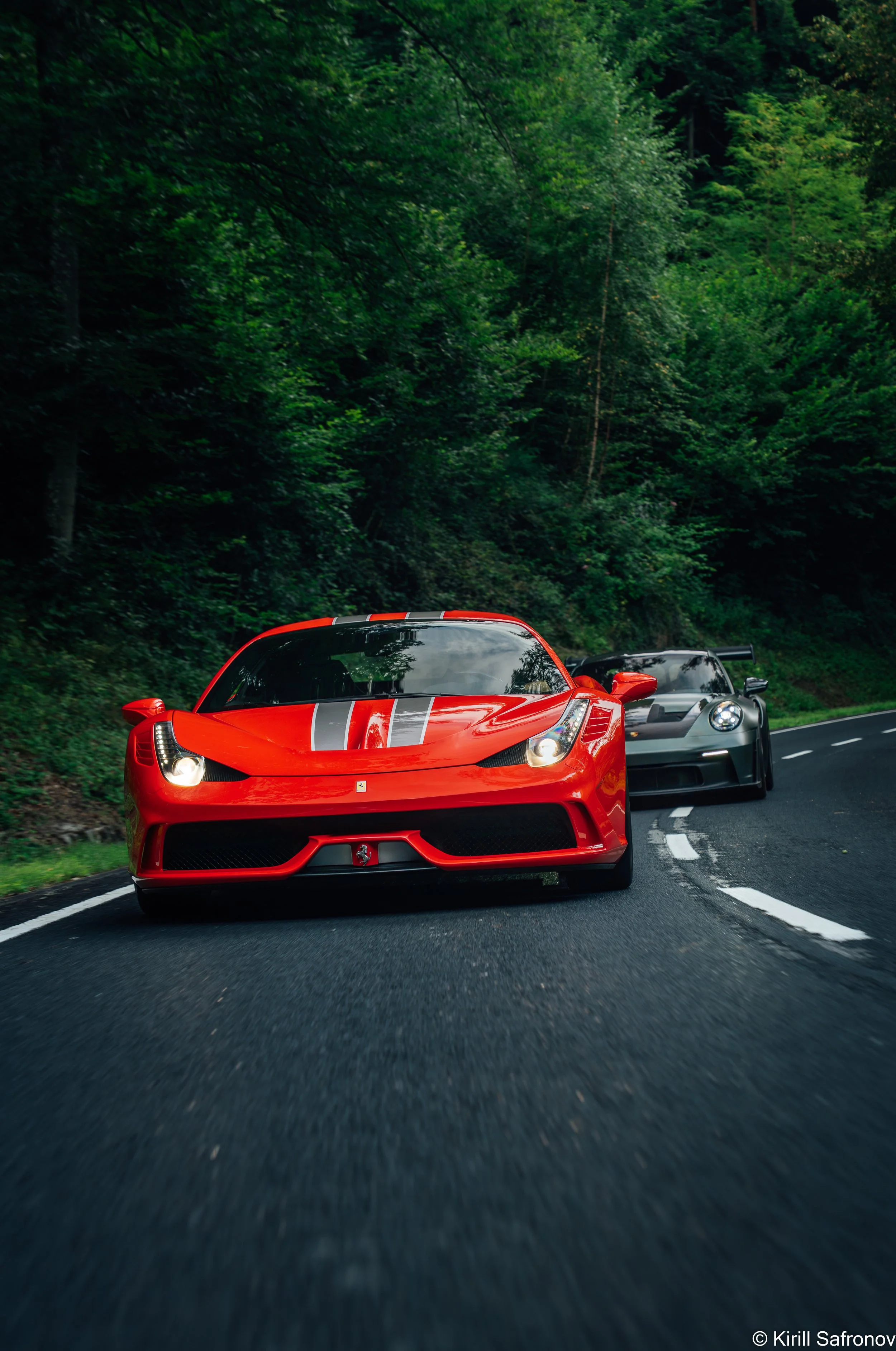 A red Ferrari 458 Speciale with silver racing stripes on the hood driving on a scenic road surrounded by dense forest.