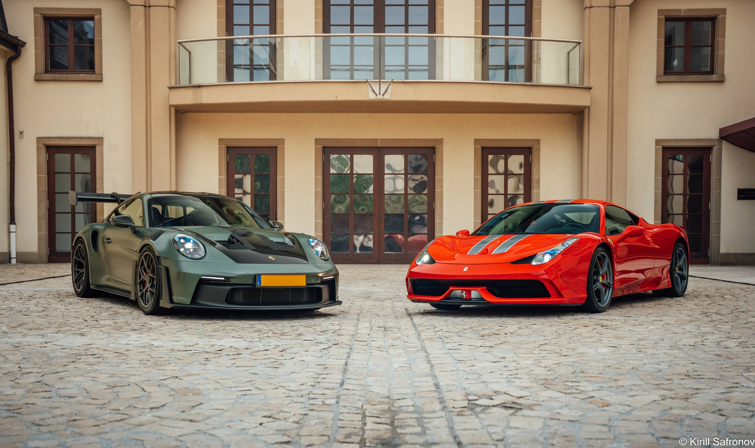 Dark green Porsche 911 GT3 RS and red Ferrari 458 Speciale parked in front of an elegant building with wooden windows and a glass balcony.