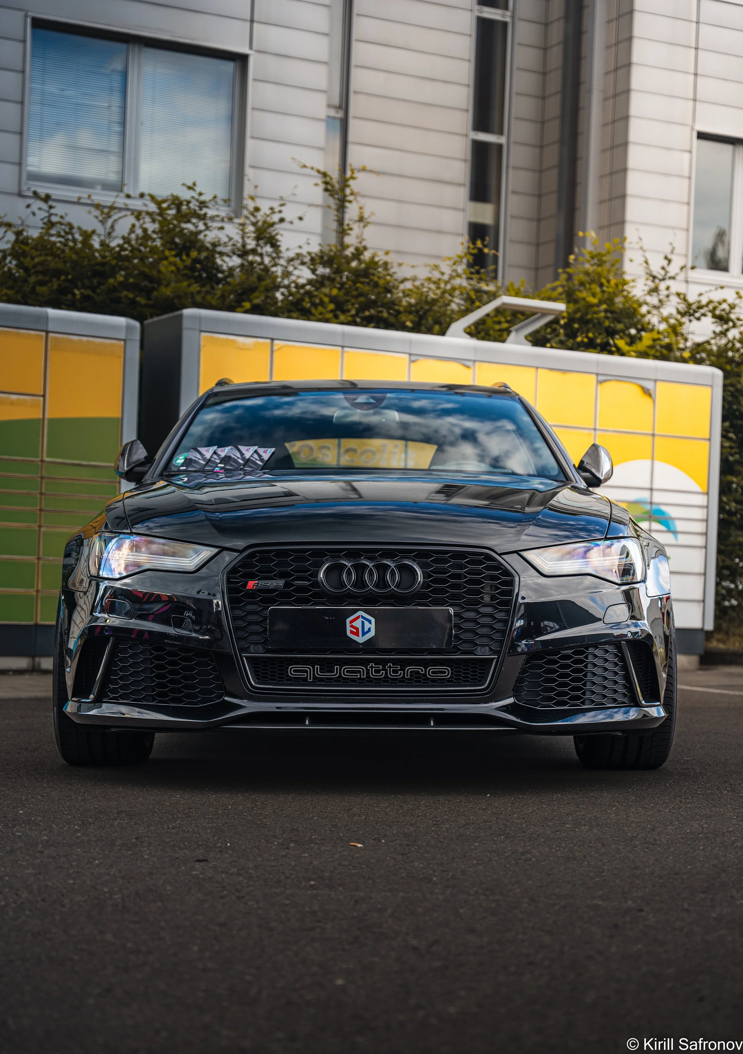 Photo of a front view black Audi quattro RS6 C7 driving past a bus stop with yellow signs and a green hedge in the background.