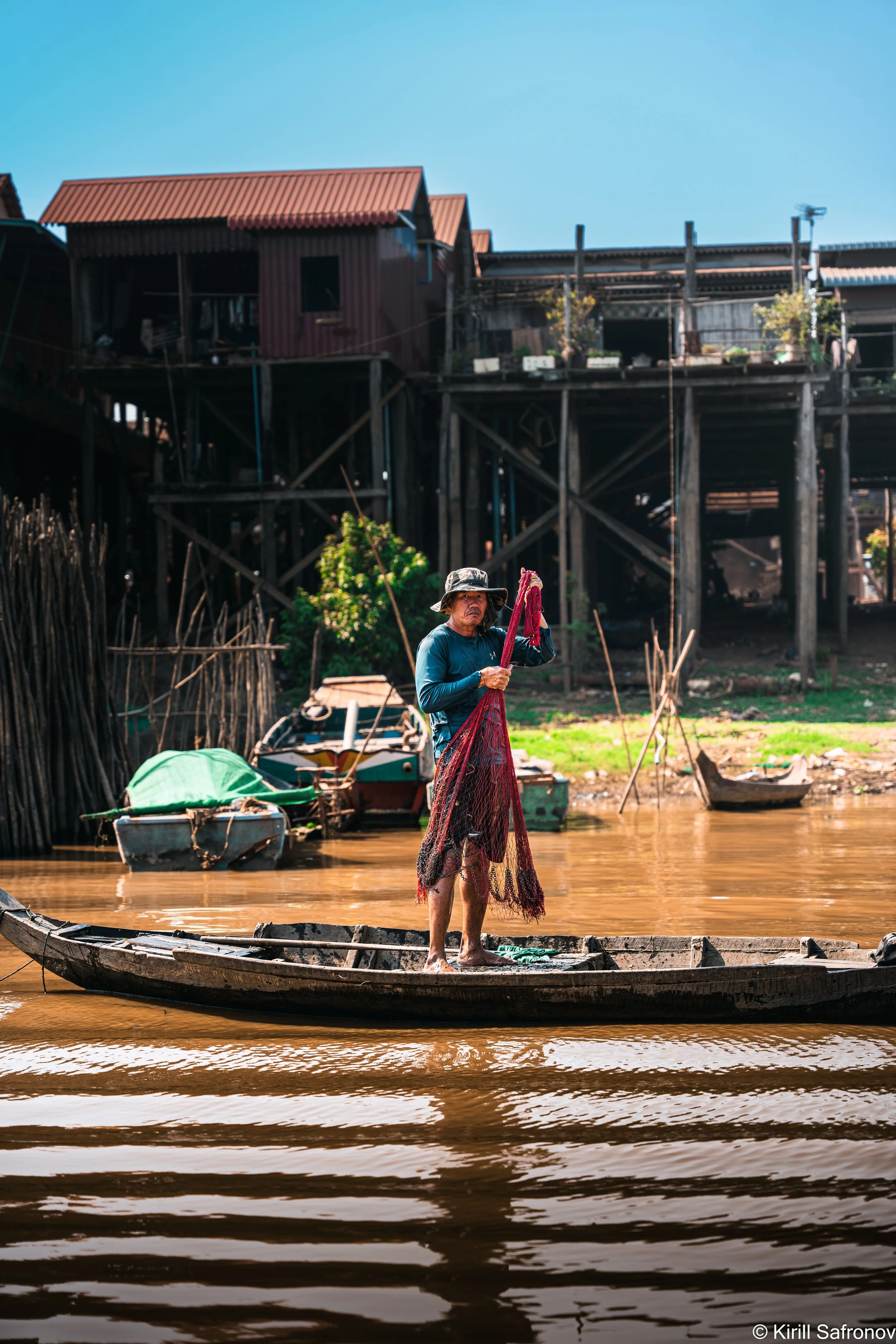 Photo of a fisherman standing on a small wooden boat by the water, holding a red fishing net near traditional wooden stilt houses under a clear blue sky.