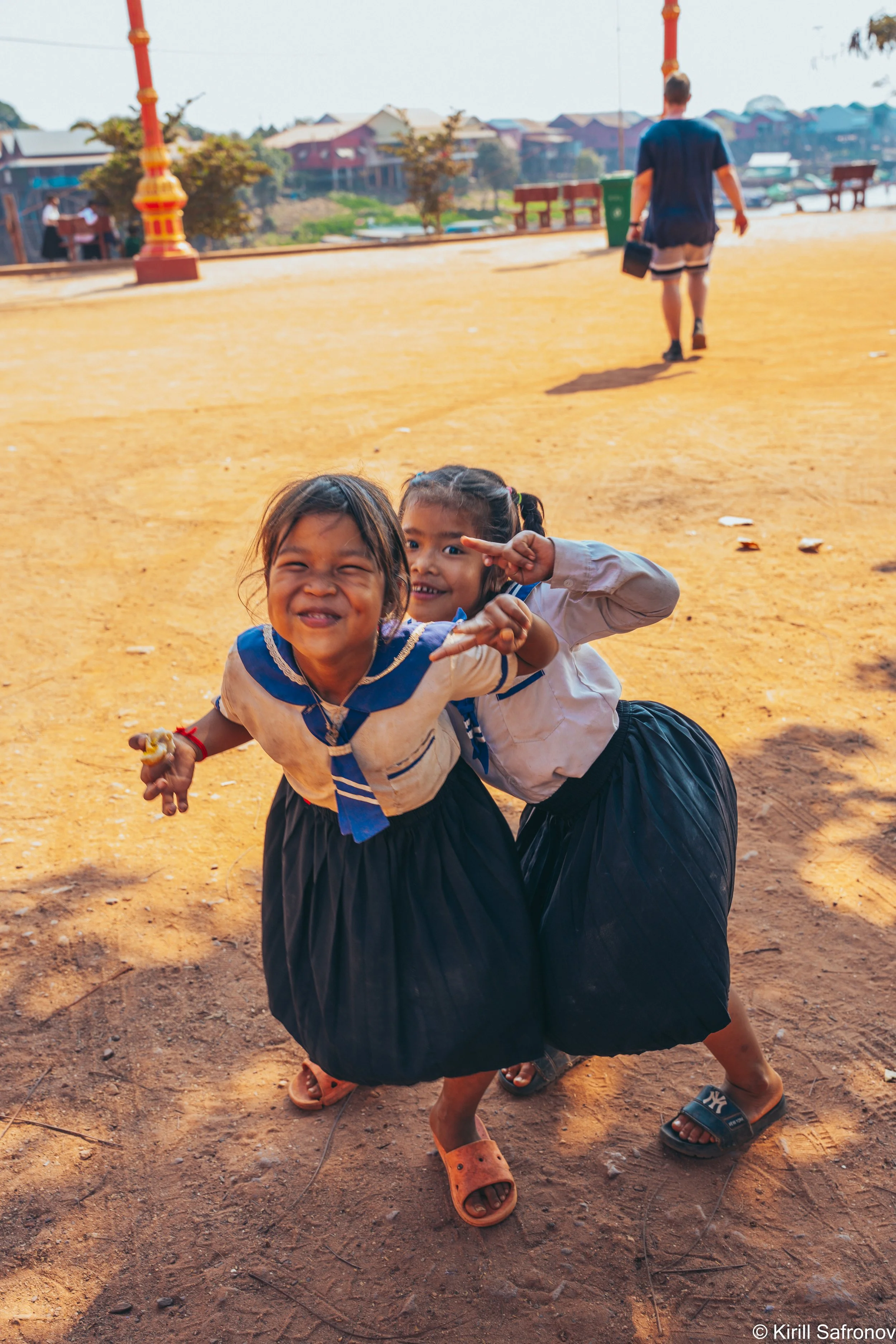 Photo of two young girls in school uniforms smiling and posing outdoors, with another child walking in the background on a dirt ground in an urban neighborhood.