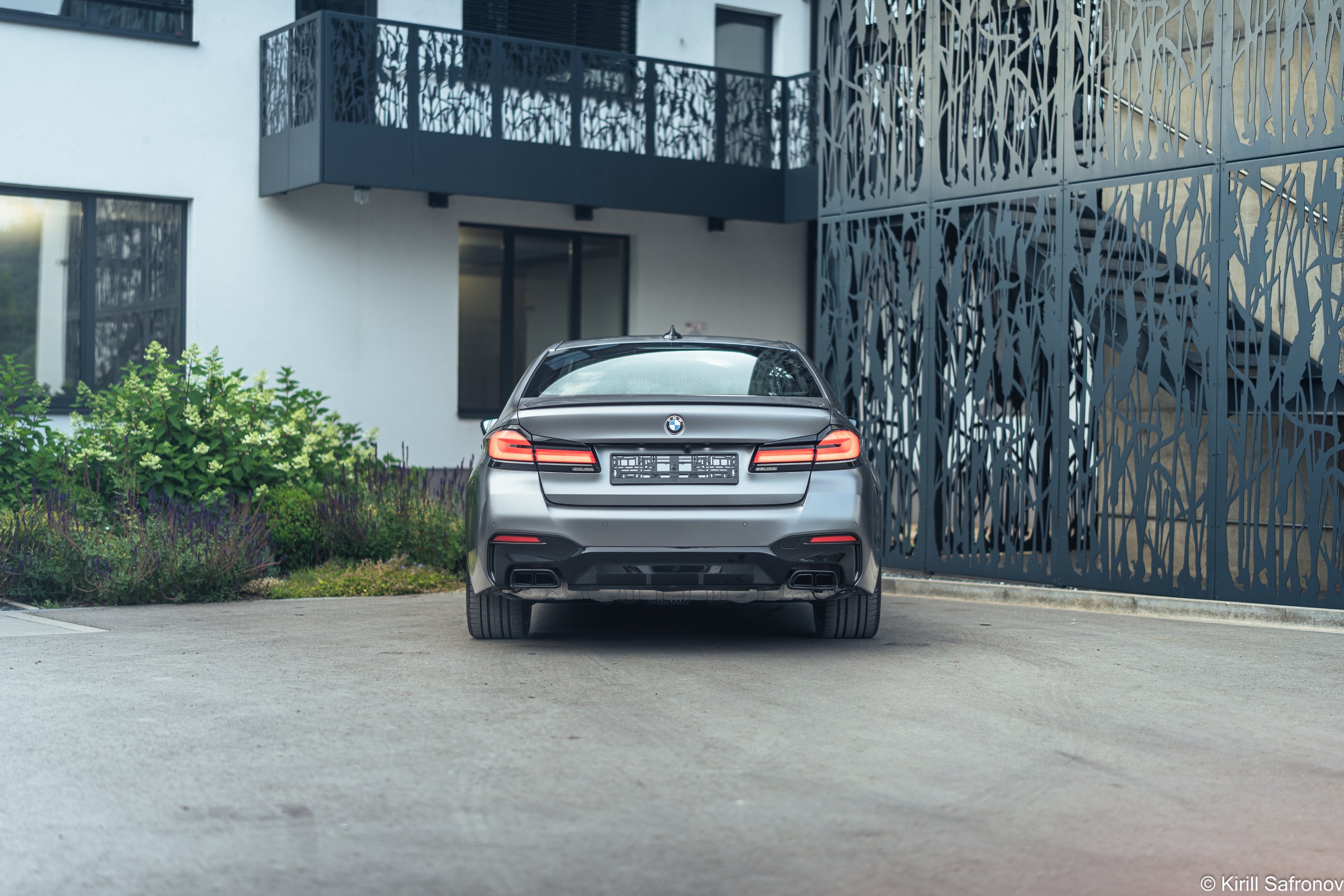 rear view Photo of a grey BMW M550i luxury sedan parked outside a modern architectural building with a decorative metal facade.