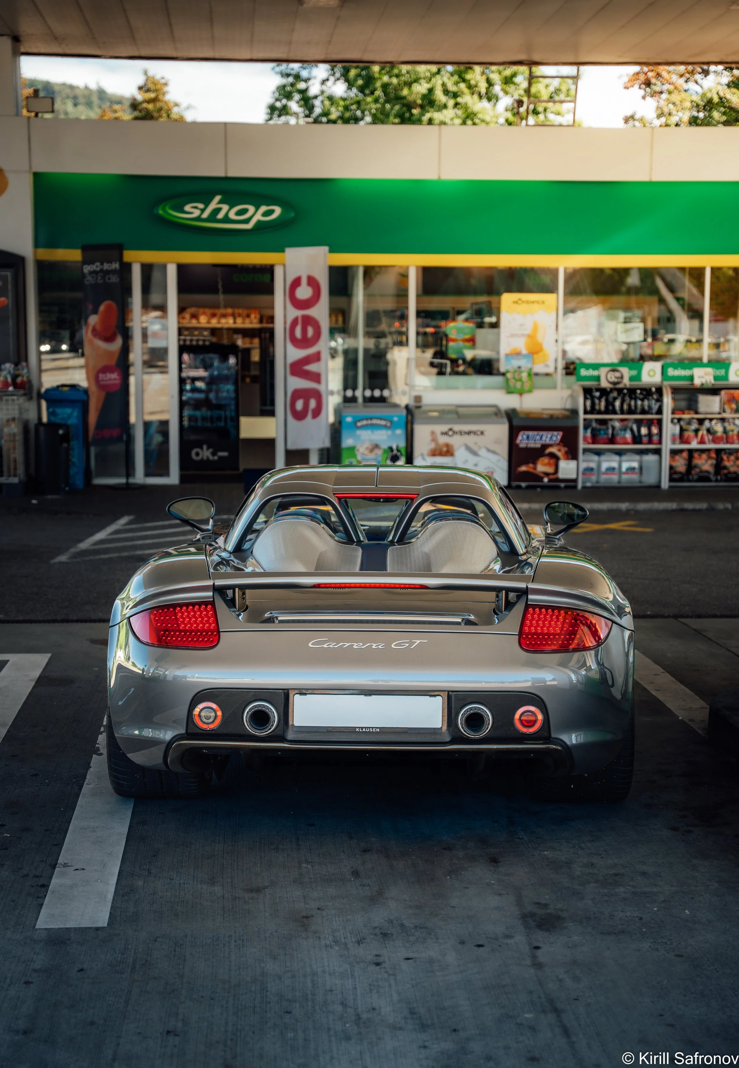 Photo of a silver Porsche Carrera GT sports car parked under a building near a supermarket entrance with a green shop sign in the background.
