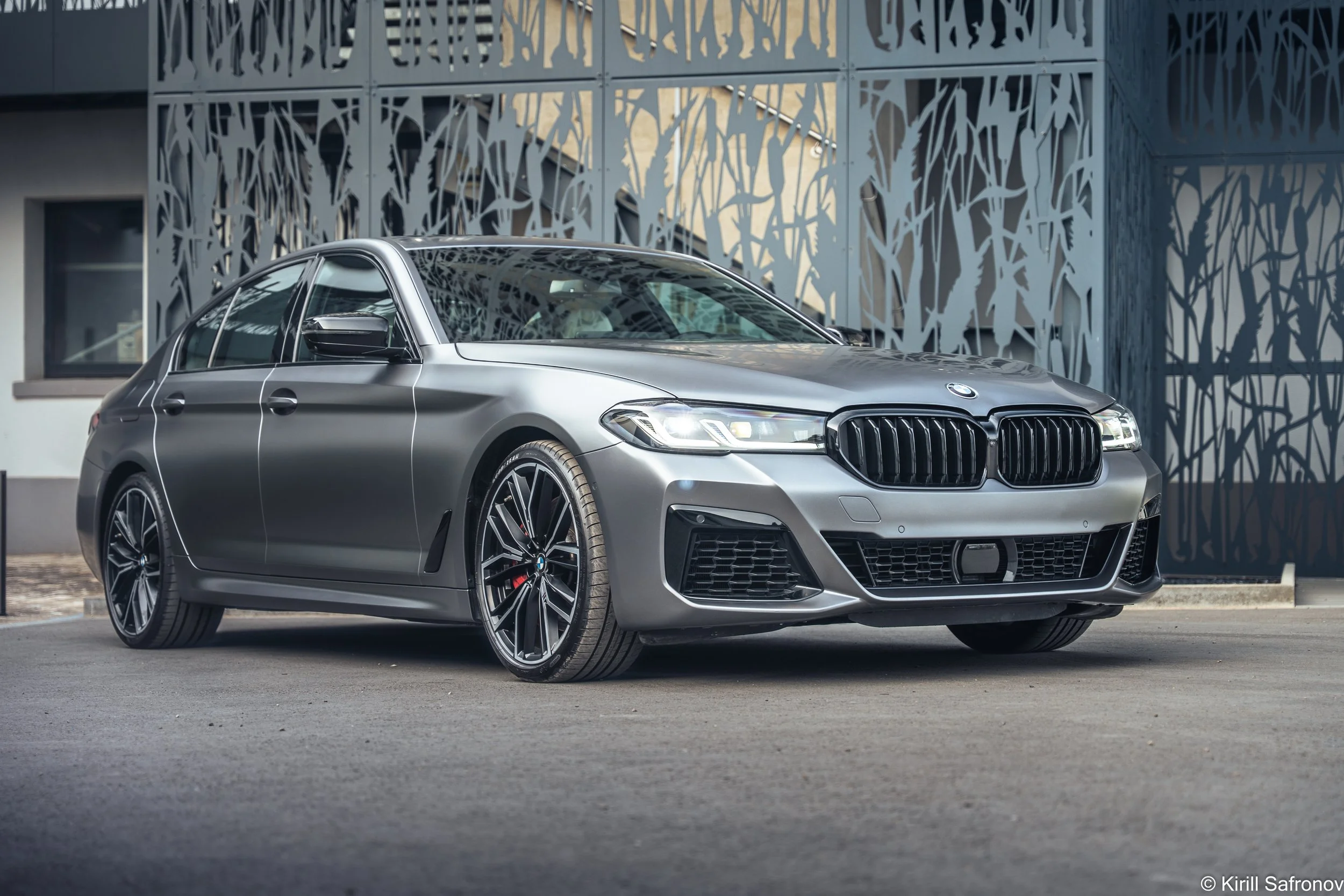 Photo of a grey BMW M550i luxury sedan parked outside a modern architectural building with a decorative metal facade.