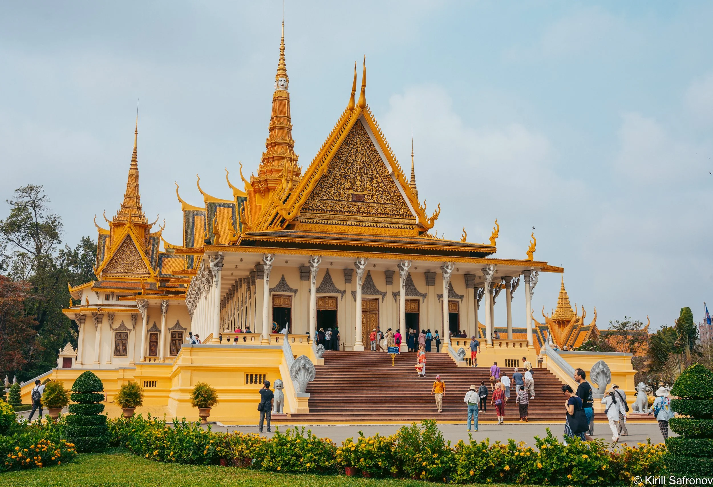 Photo of the Royal Palace in Phnom Penh, Cambodia, featuring golden ornate architecture with multiple pointed roofs, visitors climbing the stairs, and a beautifully maintained garden with shrubs in the foreground.