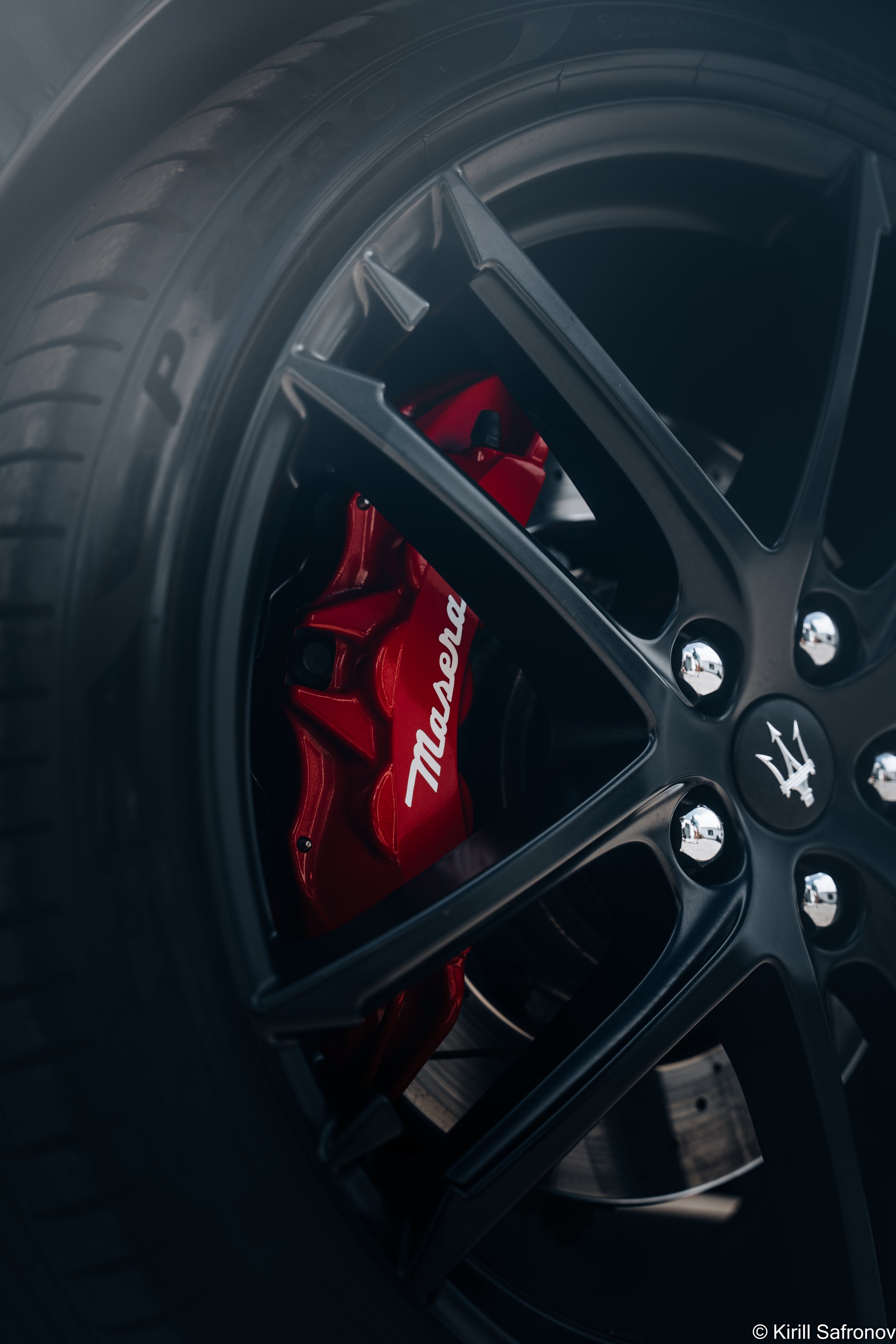 Photo of a Maserati car wheel close-up with a red disc brake caliper, white Maserati logo, alloy rim, and black rubber performance tire.