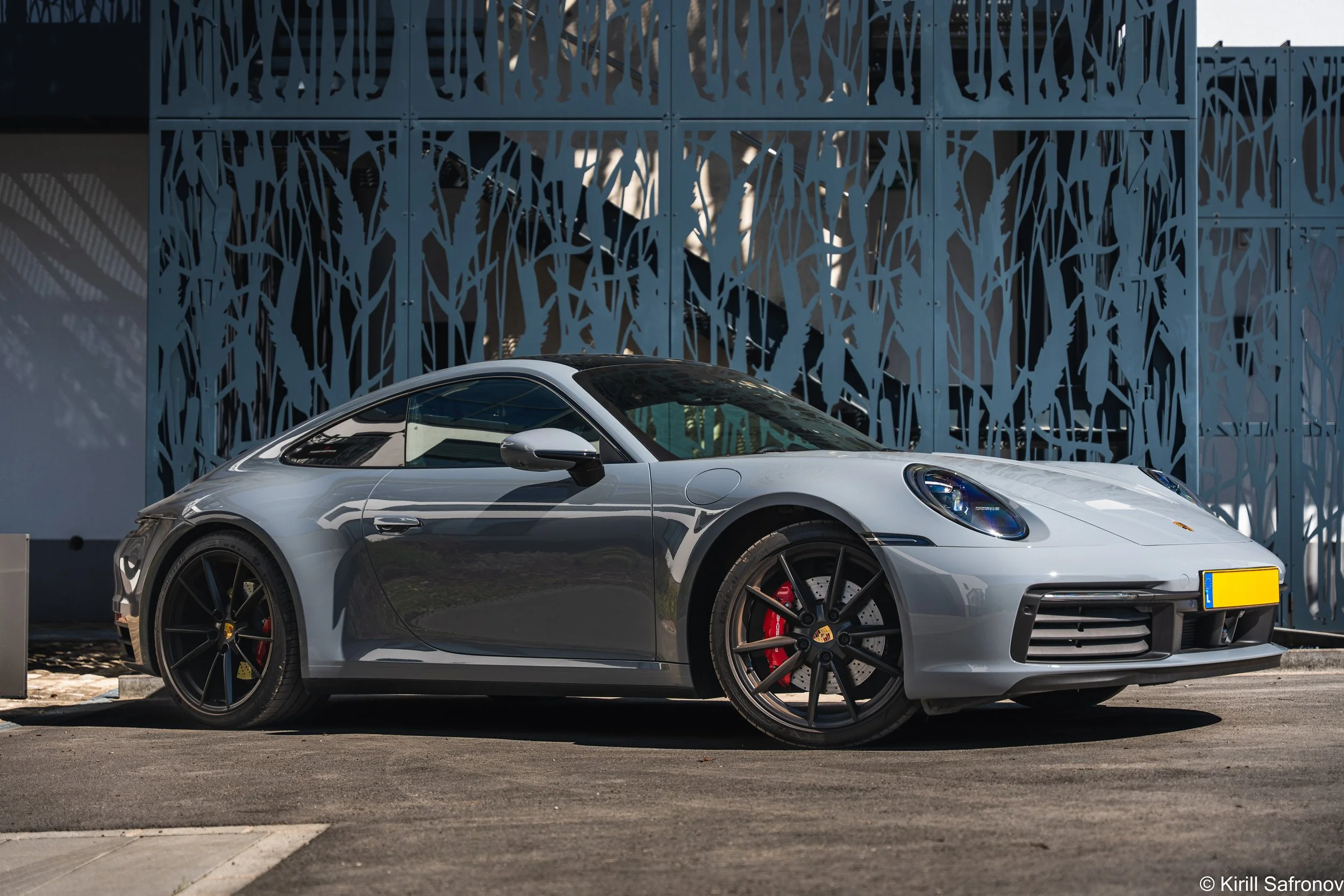A gray Porsche 911 Carrera 4S sports car parked outside a modern architectural building with a perforated metal facade featuring tree-branch patterns, photographed under bright sunny skies.