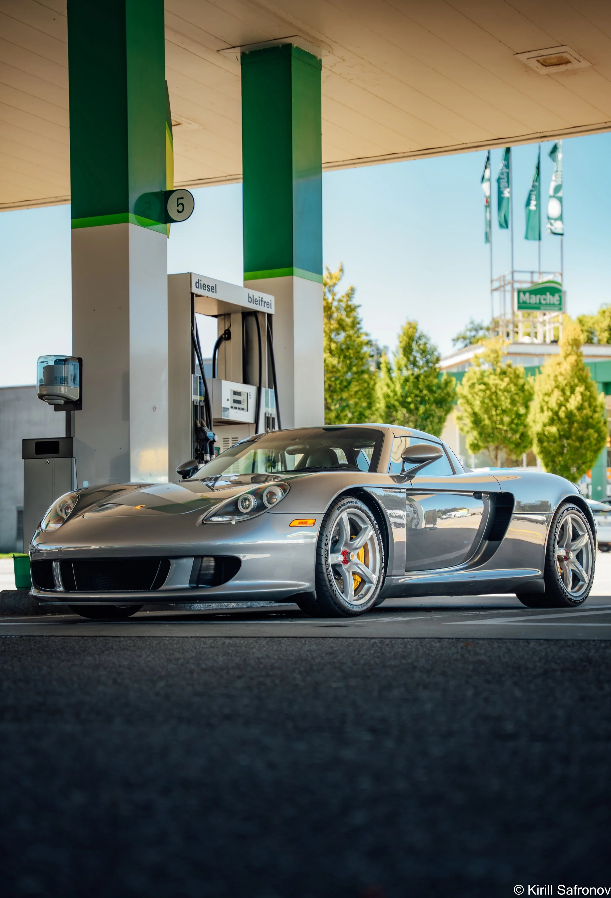 A photo of a silver Porsche Carrera GT sports car parked at a gas station, with fuel pumps, French signage, green trees, and a clear blue sky in the background.