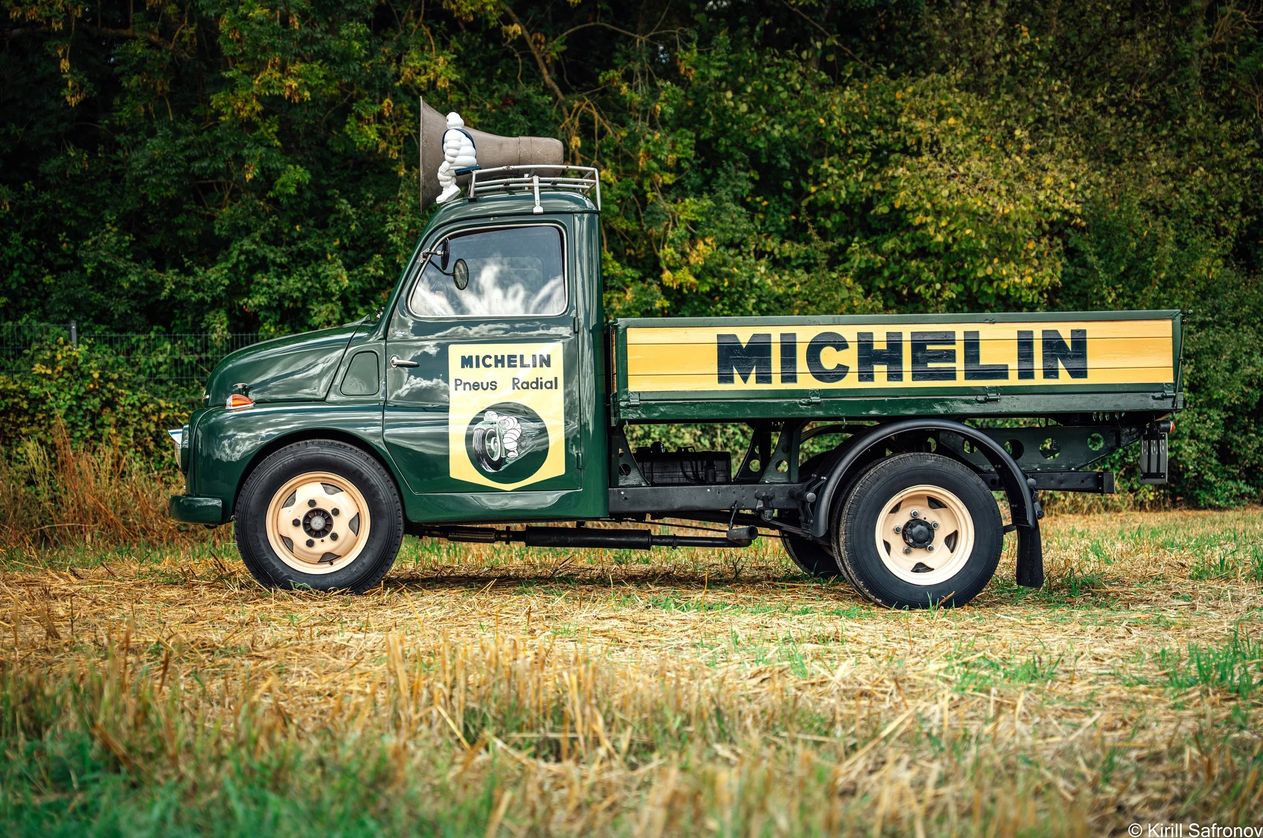Photo of a dark green 1995 Fiat 615 vintage truck with a Michelin advertising panel, roof loudspeaker, and Michelin Man mascot, parked outdoors in a natural landscape with trees and dry grass.