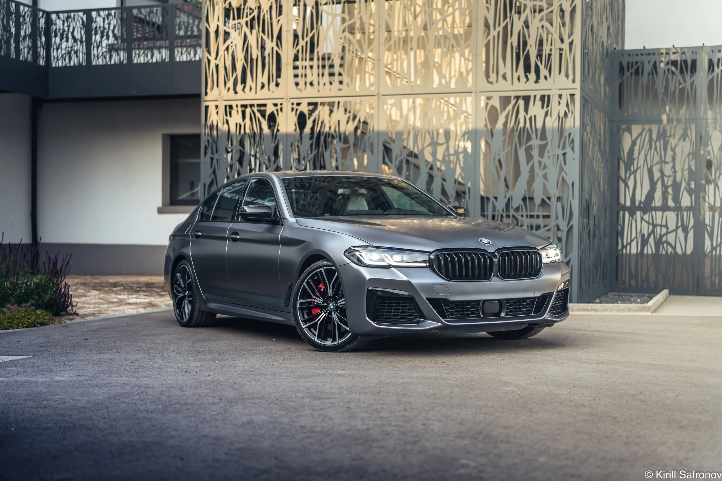 Photo of a grey BMW M550i luxury sedan parked outside a modern architectural building with a decorative metal facade.