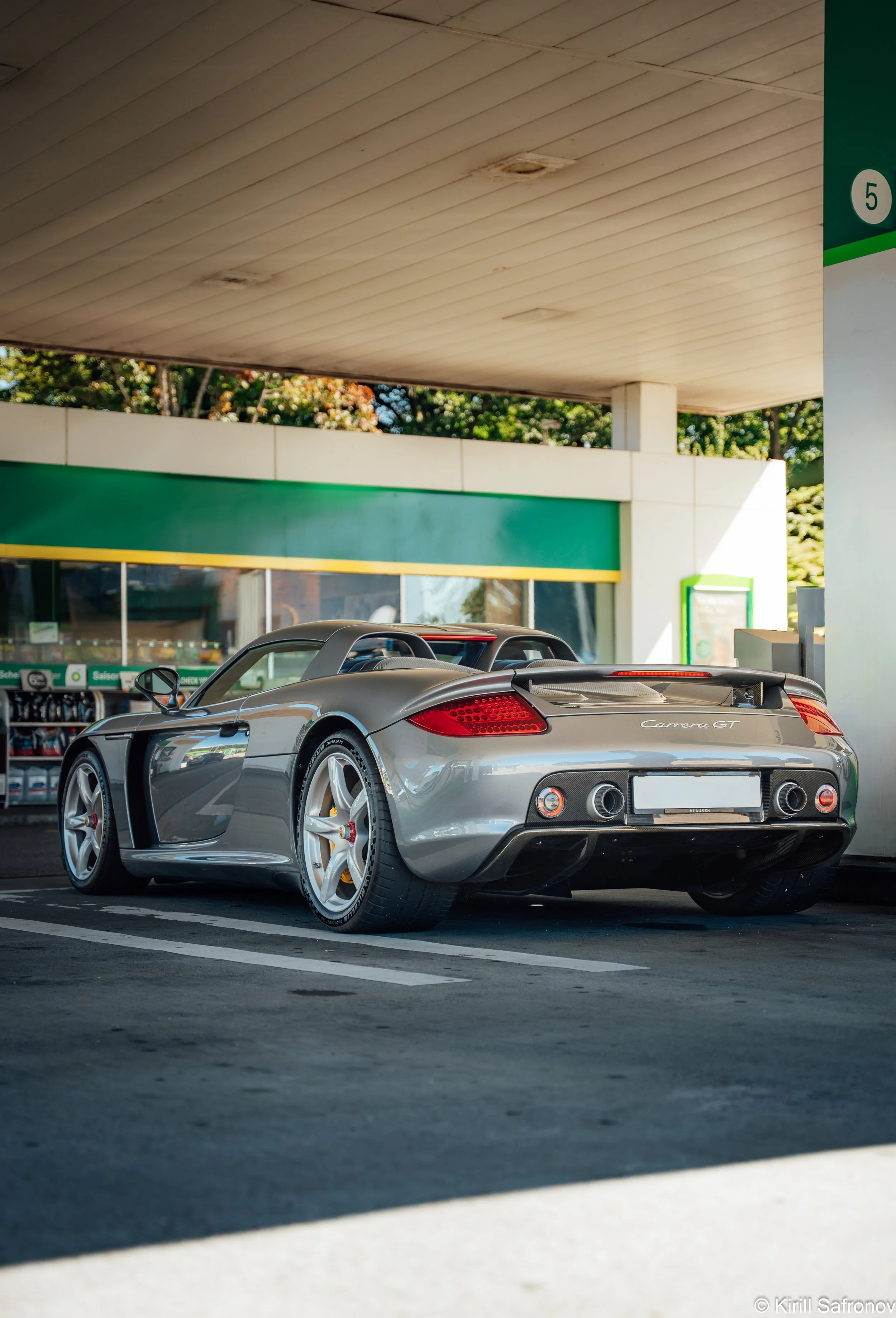 A gray Porsche Carrera GT sports car photo parked in front of a gas station, with a modern building in the background and a wooden canopy overhead during daylight.