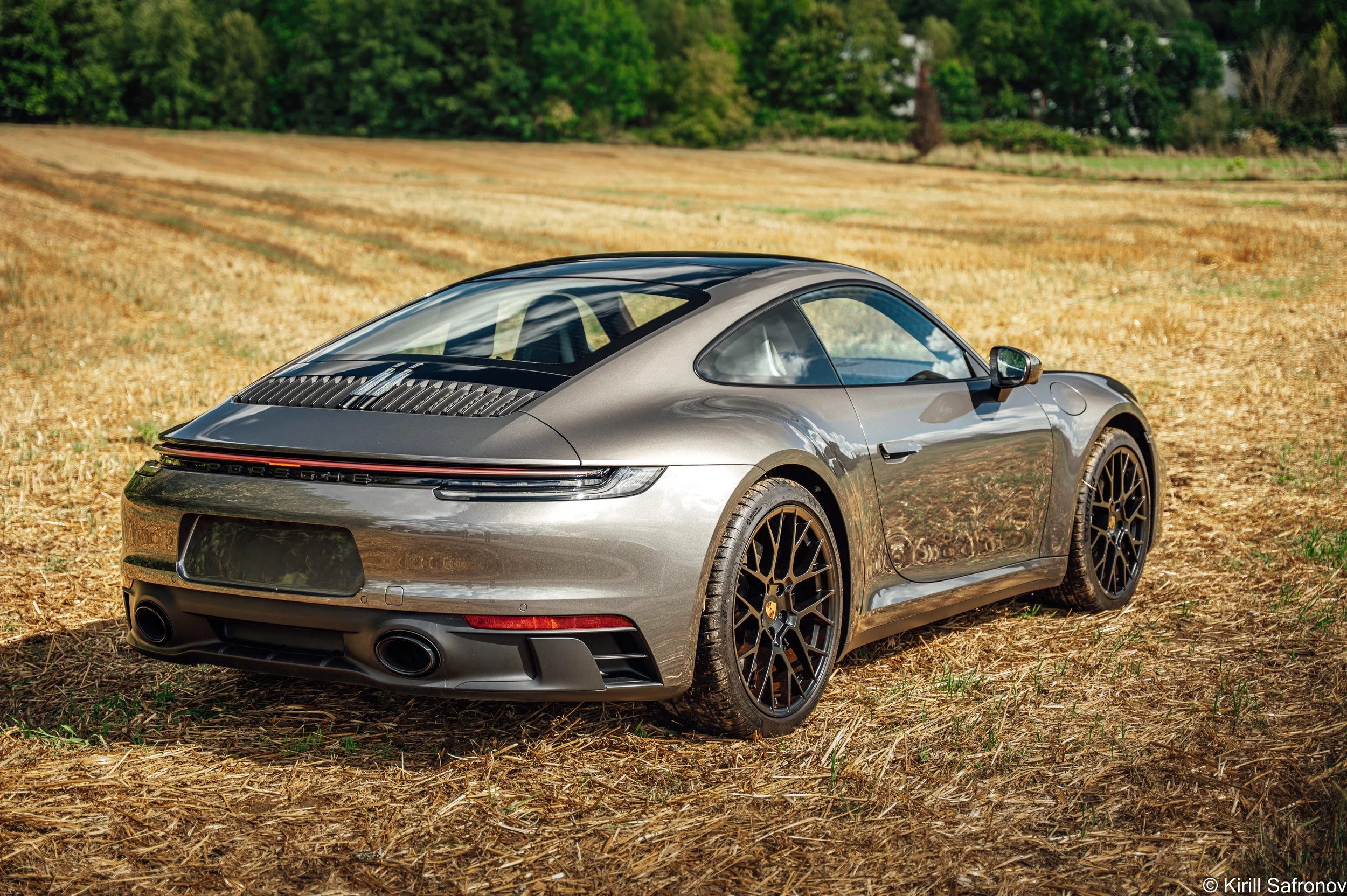 A metallic champagne Porsche 911 Carrera GTS parked in an open field, surrounded by nature with trees in the background.