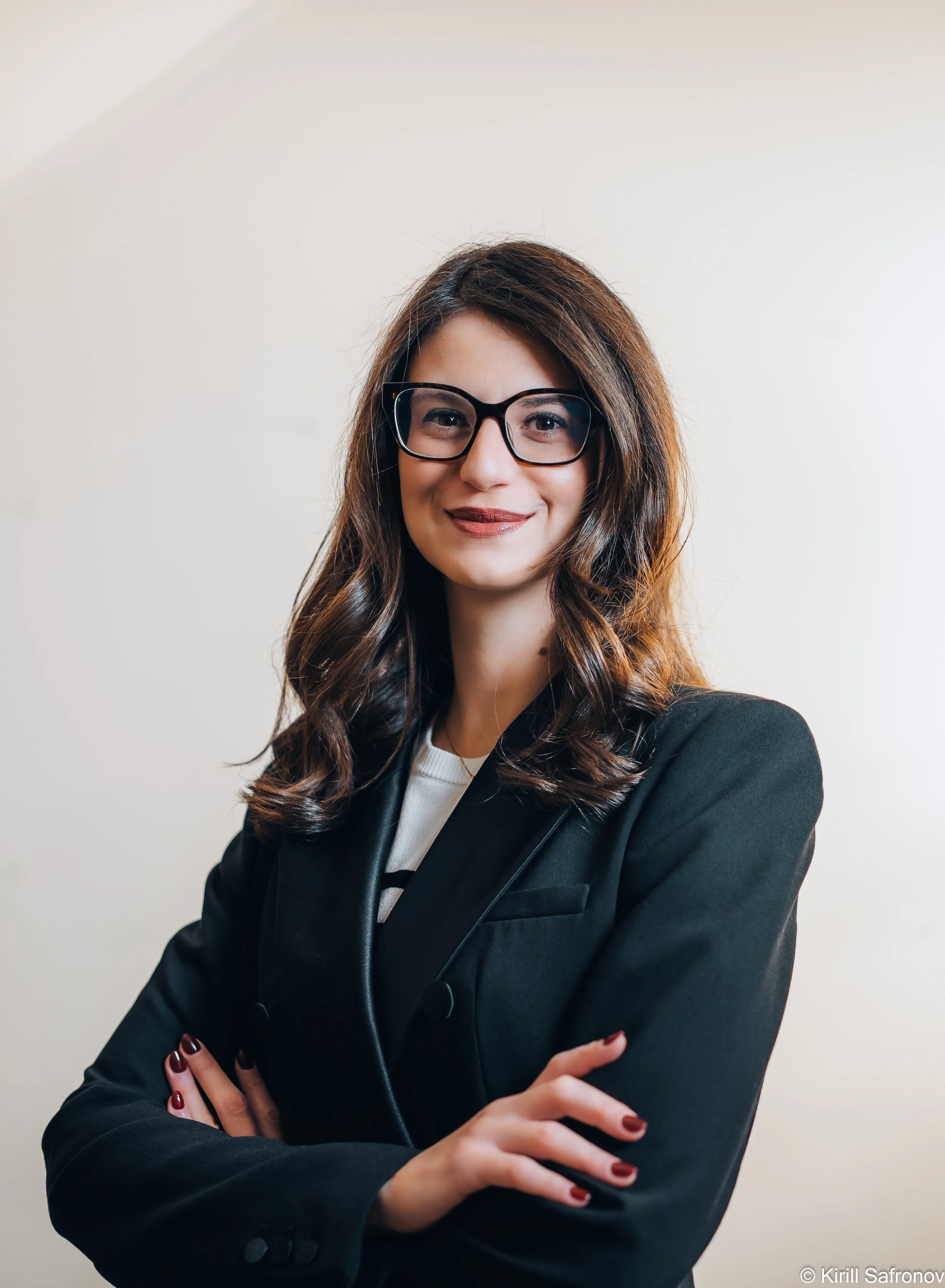 Photo of a confident smiling businesswoman wearing black sunglasses and a black blazer, standing with crossed arms in a professional corporate setting.
