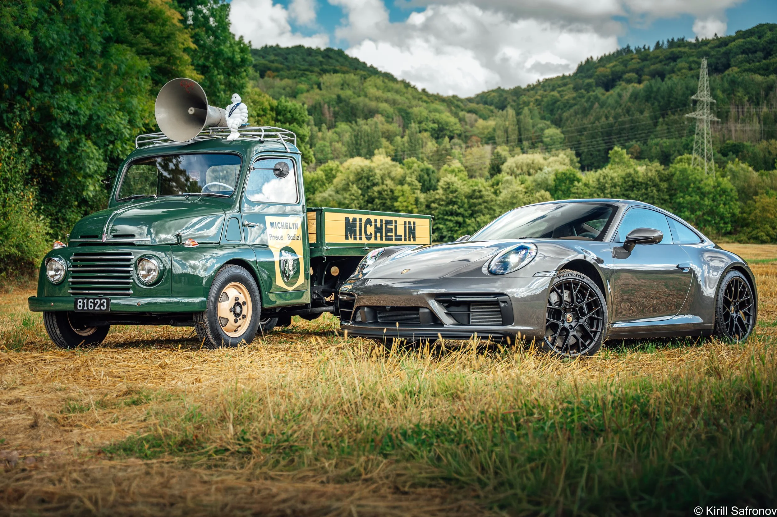 Automotive photo featuring a champagne Porsche 911 Carrera GTS positioned in front of a dark green 1995 Fiat 615 vintage truck with a Michelin advertising sign. Captured in a scenic countryside landscape with rolling green hills and a cloudy sky, thi