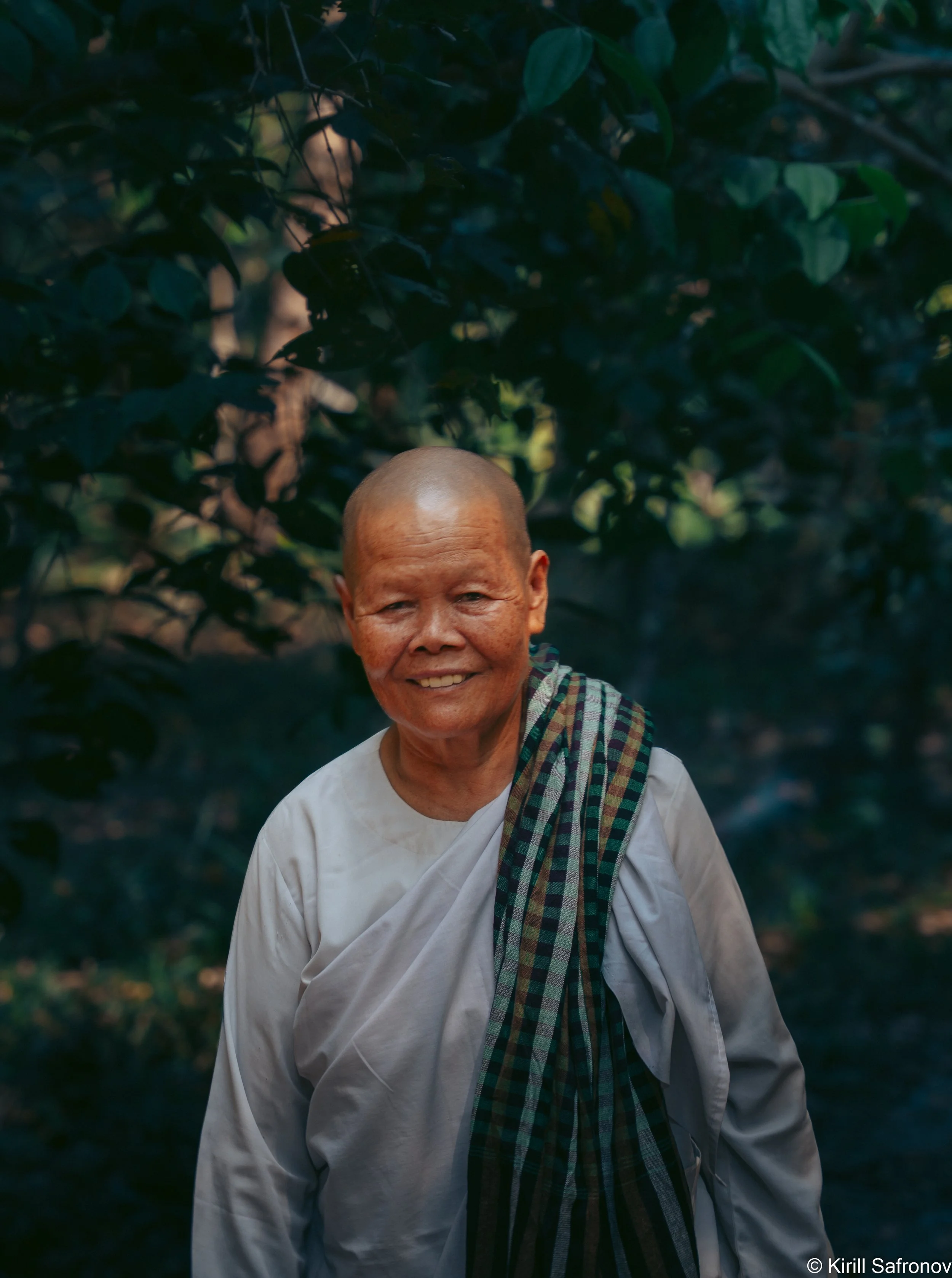 Photo of a Cambodian Buddhist nun with a shaved head, wearing a white shirt and a tartan cloth draped over her shoulder, smiling gently in a lush green forest setting.