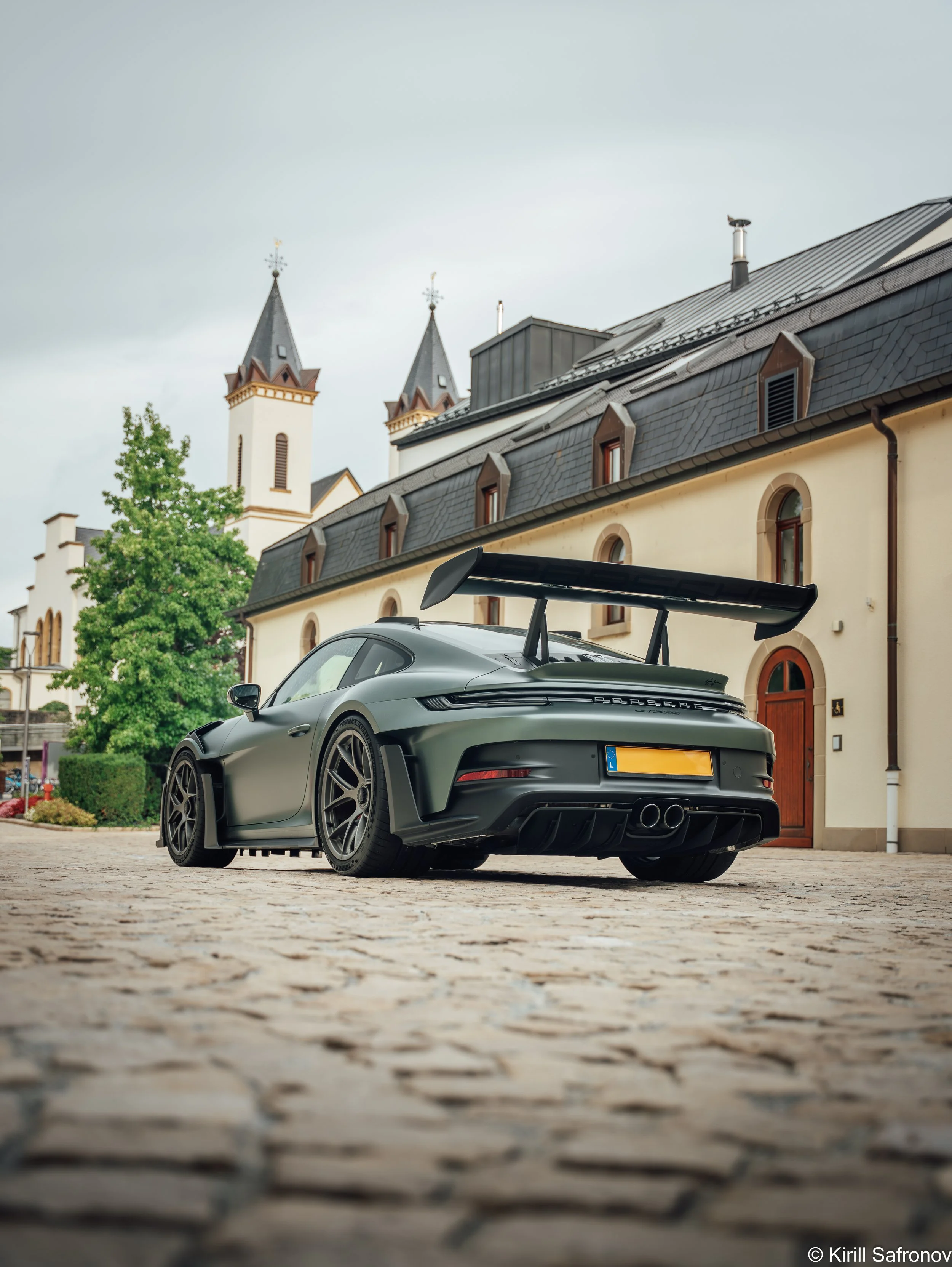 A dark green Porsche 911 GT3 RS with a large rear wing parked on a cobblestone street beside historic stone buildings, with twin church towers in the background under a cloudy sky.