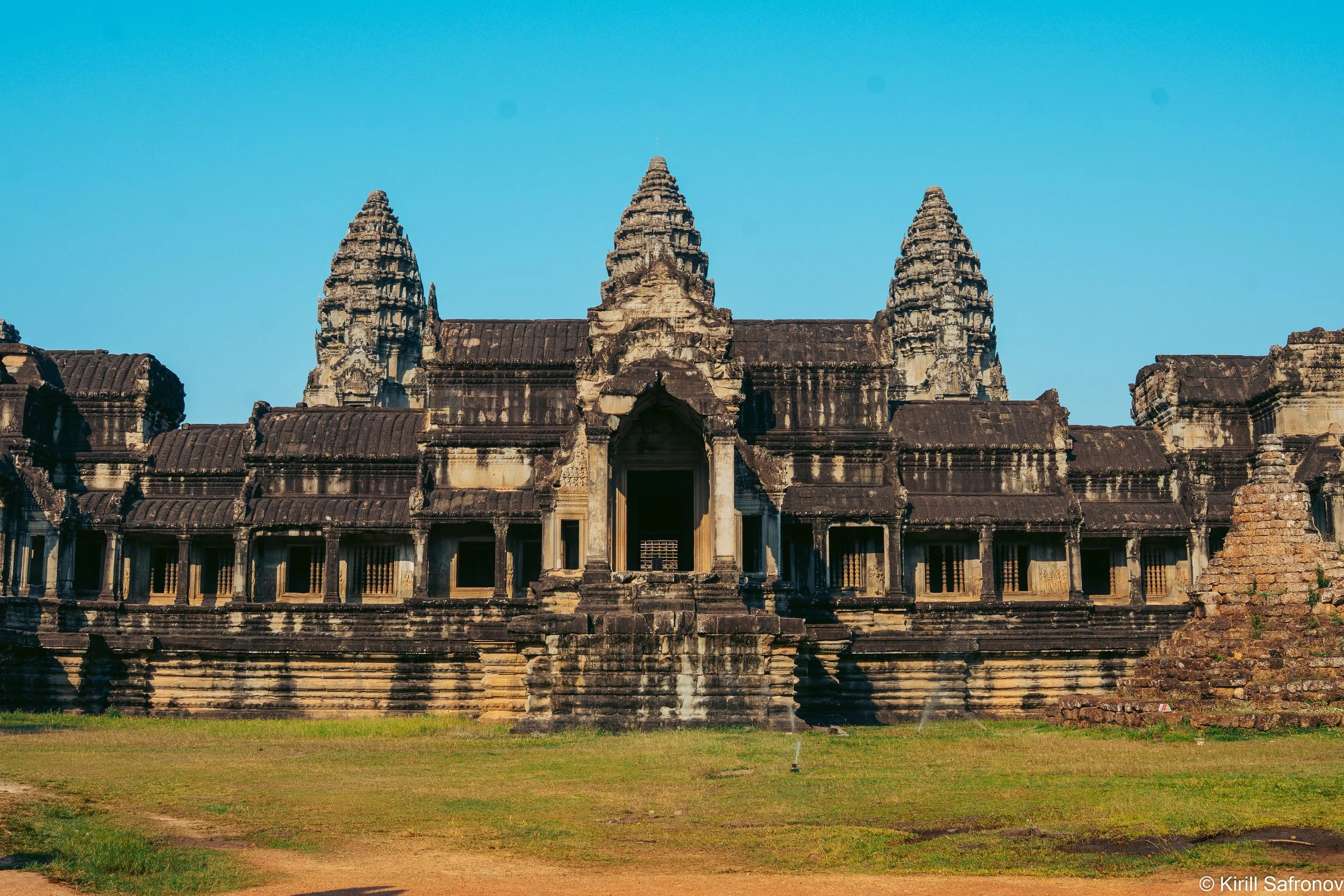 Photo of an ancient Khmer temple with three pointed stone towers, historic stone walls, and carved columns under a bright blue sky in Cambodia.