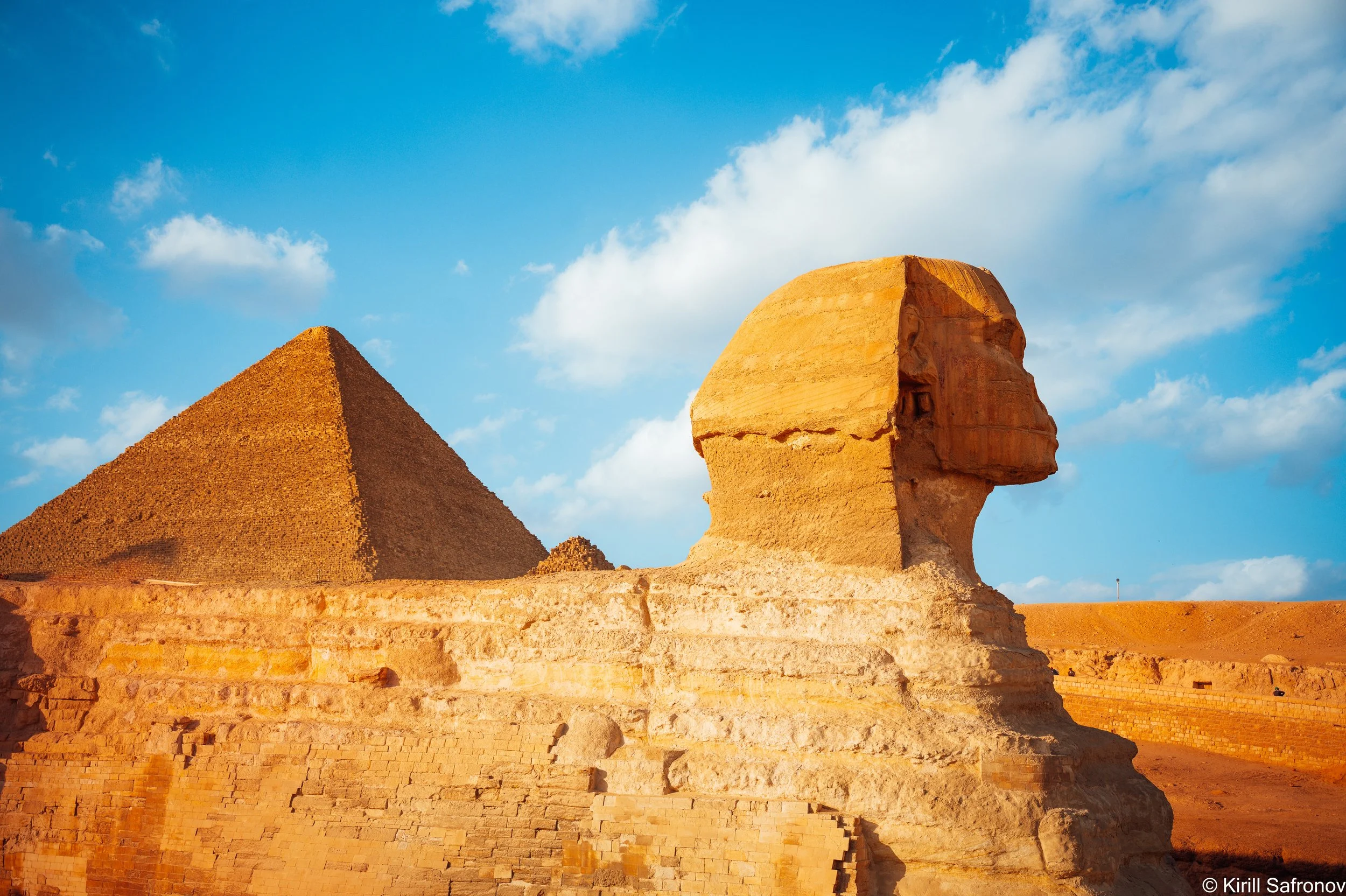 Photo of the Great Pyramid of Khufu, the ancient Egyptian pyramids of Giza, and the Sphinx under a blue sky with scattered clouds.