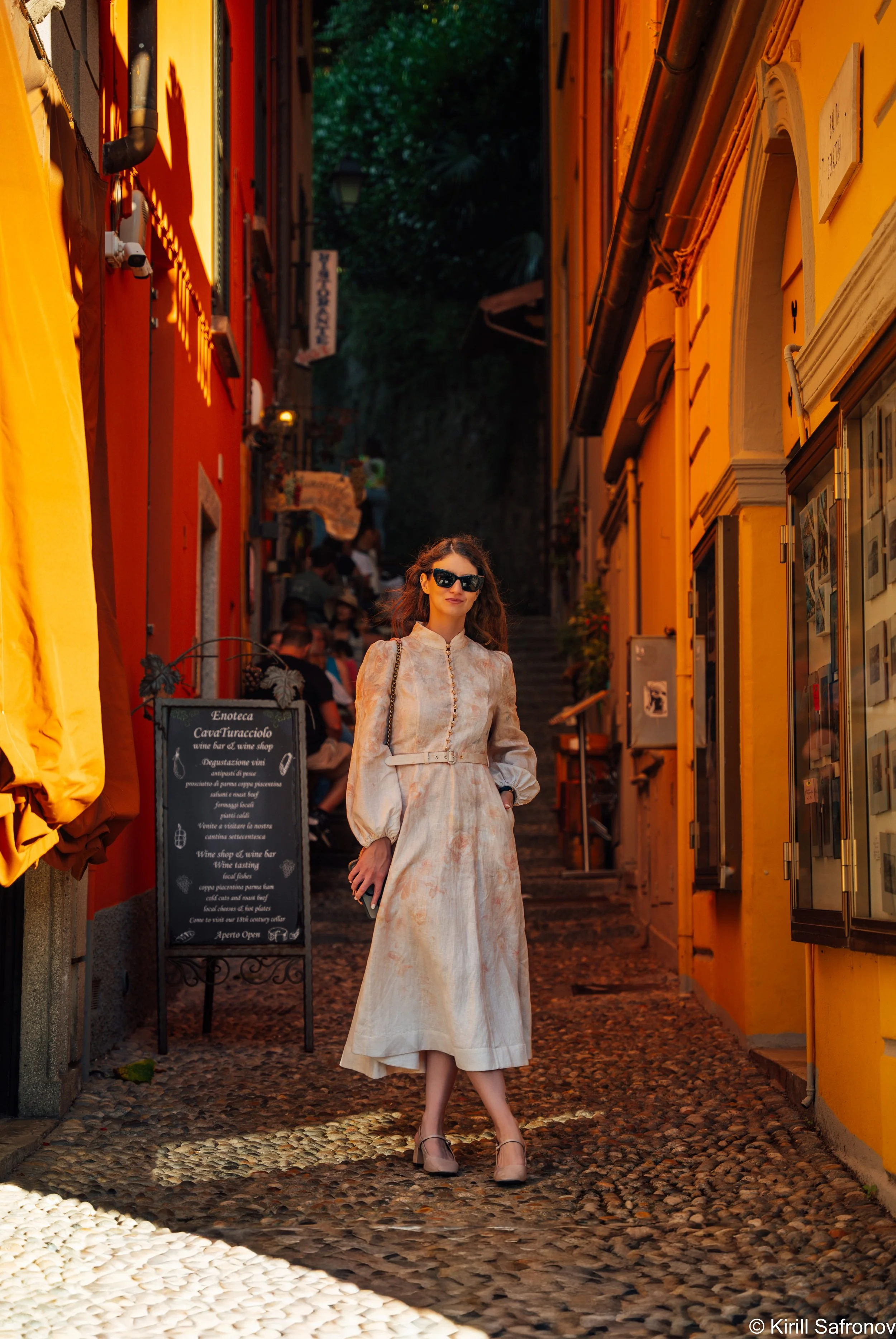 photo of an elegant woman in a beige dress, heels, and sunglasses walking along a colorful cobblestone street lined with orange and yellow buildings, with people in the background.