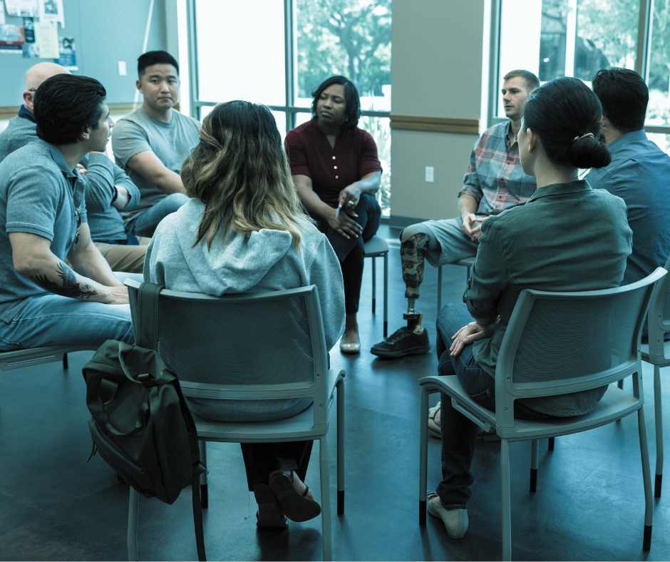 A diverse group of people seated in a circle, participating in a discussion or support group in a bright room with large windows and natural light.