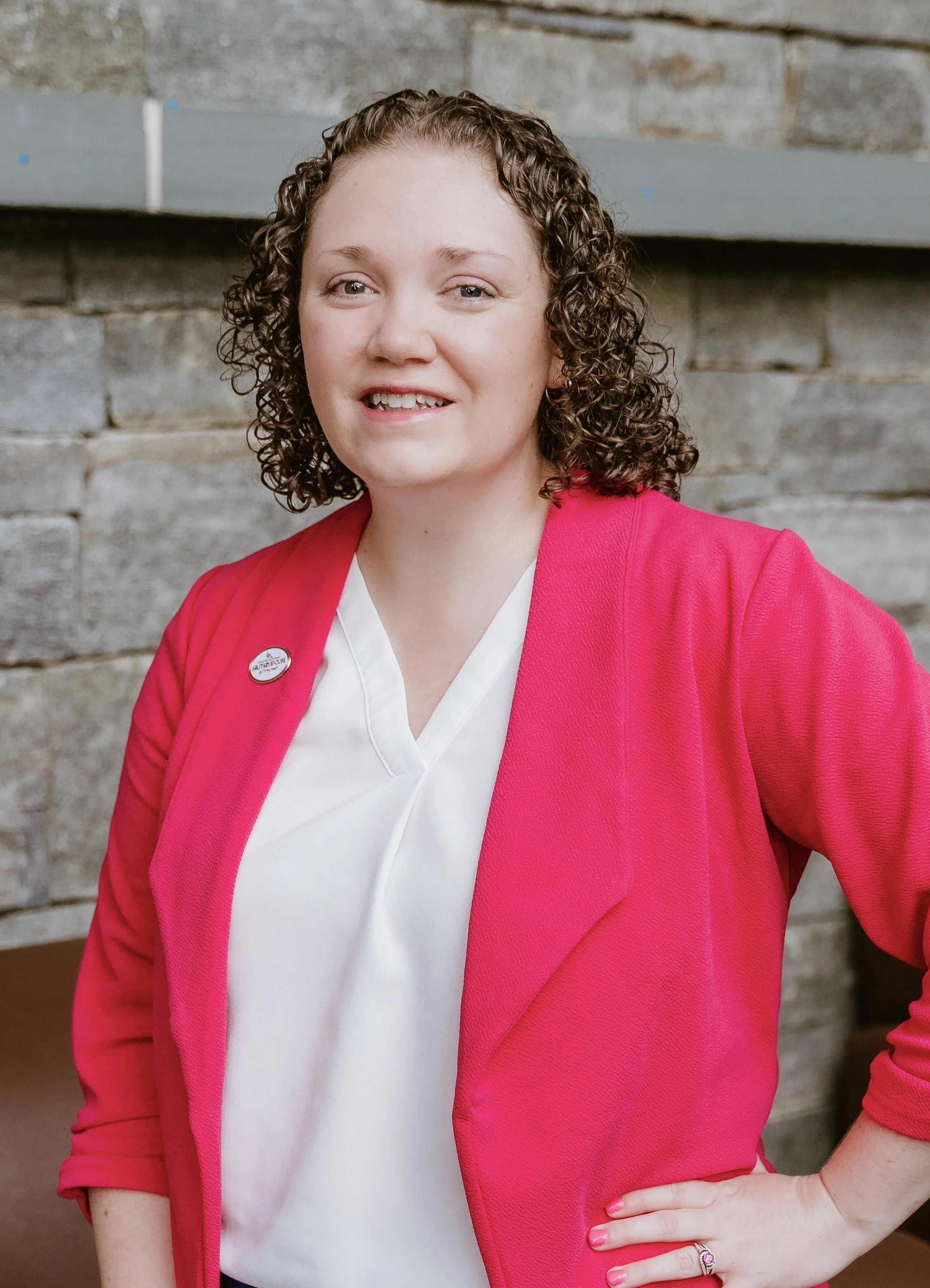 A woman with curly hair wearing a bright pink blazer over a white blouse, standing in front of a stone wall.