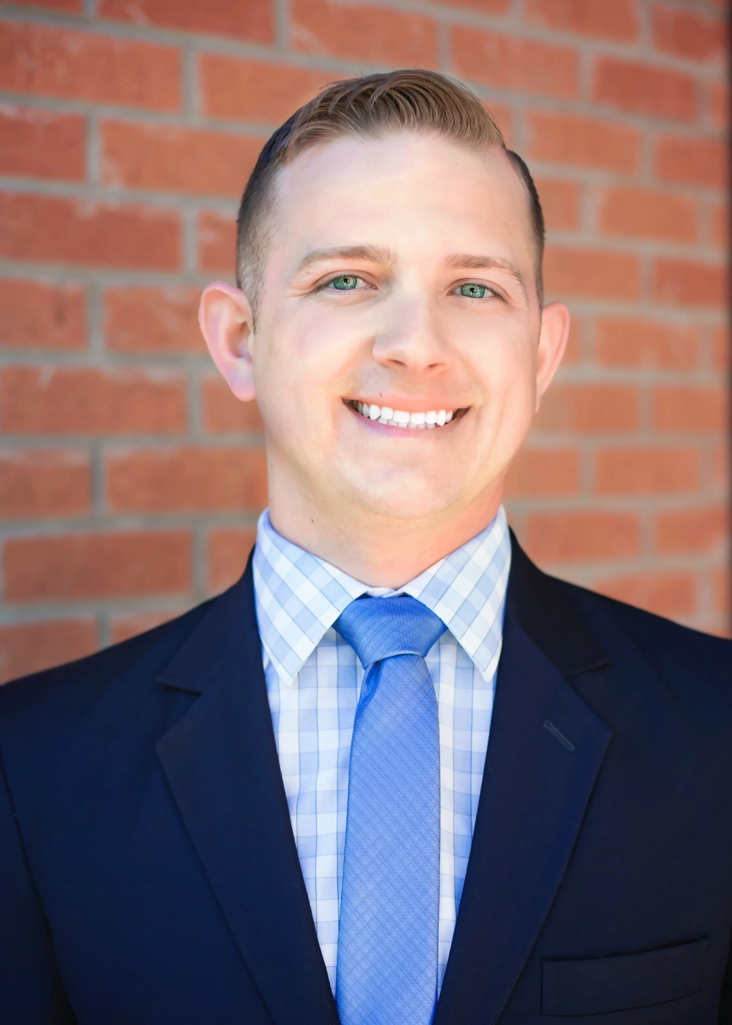 A man in a navy blue suit, white checkered shirt, and light blue tie standing in front of a brick wall, smiling and looking at the camera.
