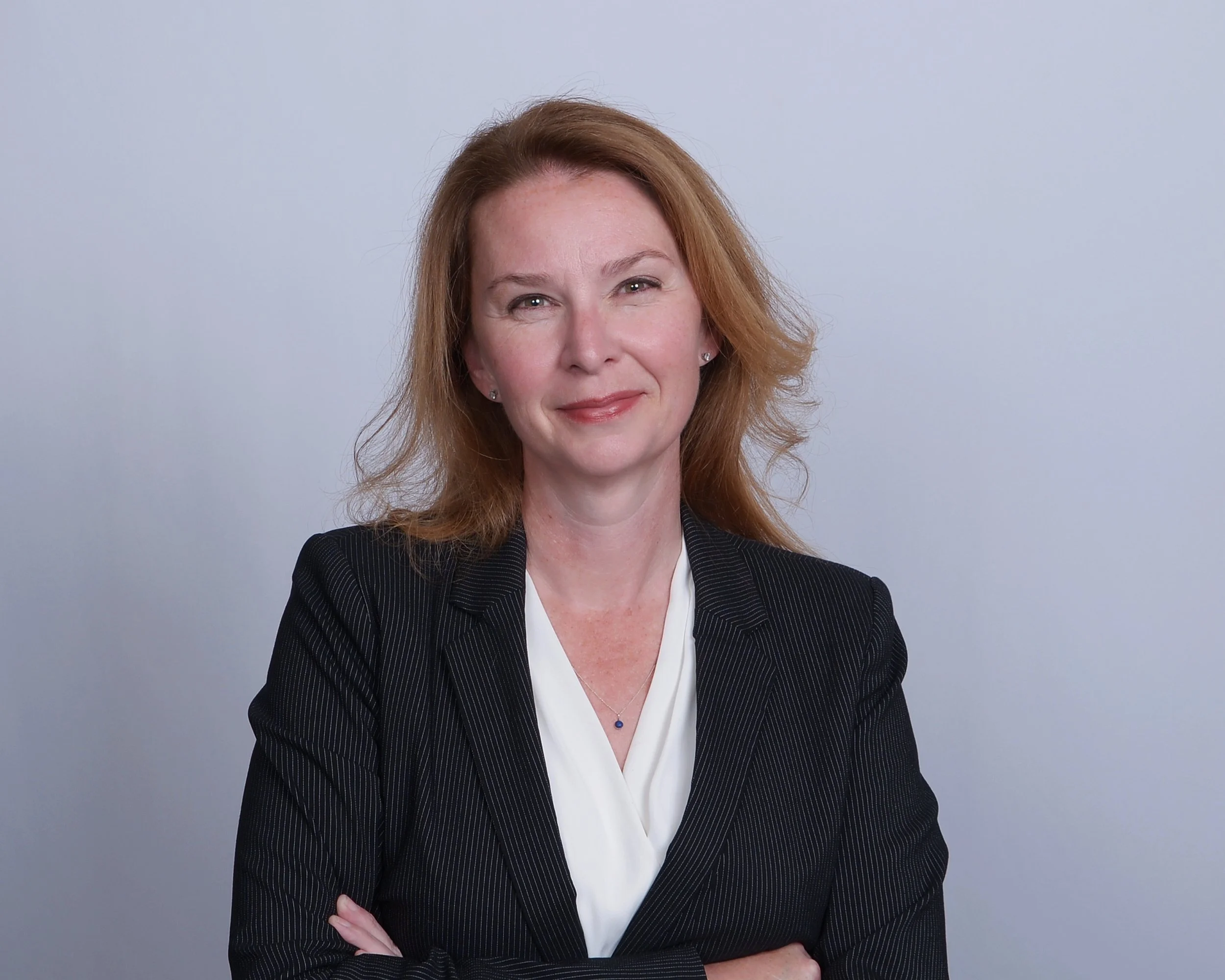 Professional woman with red hair, wearing a black pinstripe blazer and white blouse, standing with arms crossed against light gray background.